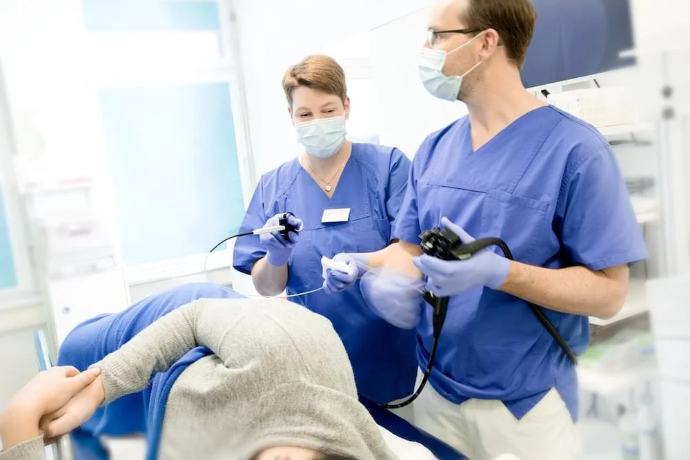 Zwei medizinische Fachkräfte in blauer Arbeitskleidung und Masken führen eine Untersuchung an einer liegenden Person durch, die auf einem Krankenbett liegt.