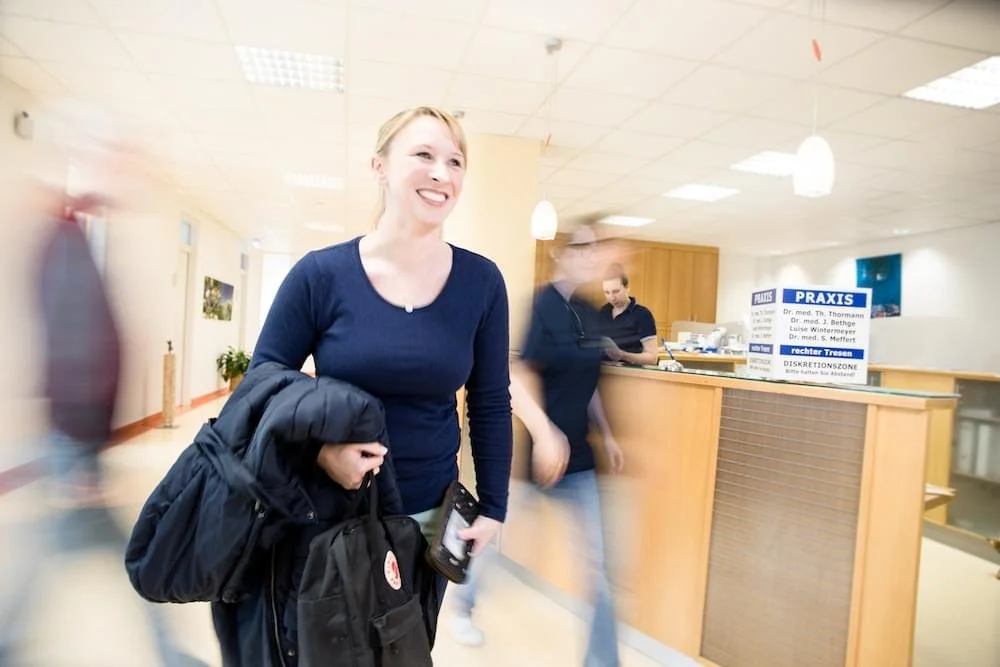 Eine lachende Frau steht an einer Rezeption in einer Arztpraxis, umgeben von anderen Personen.