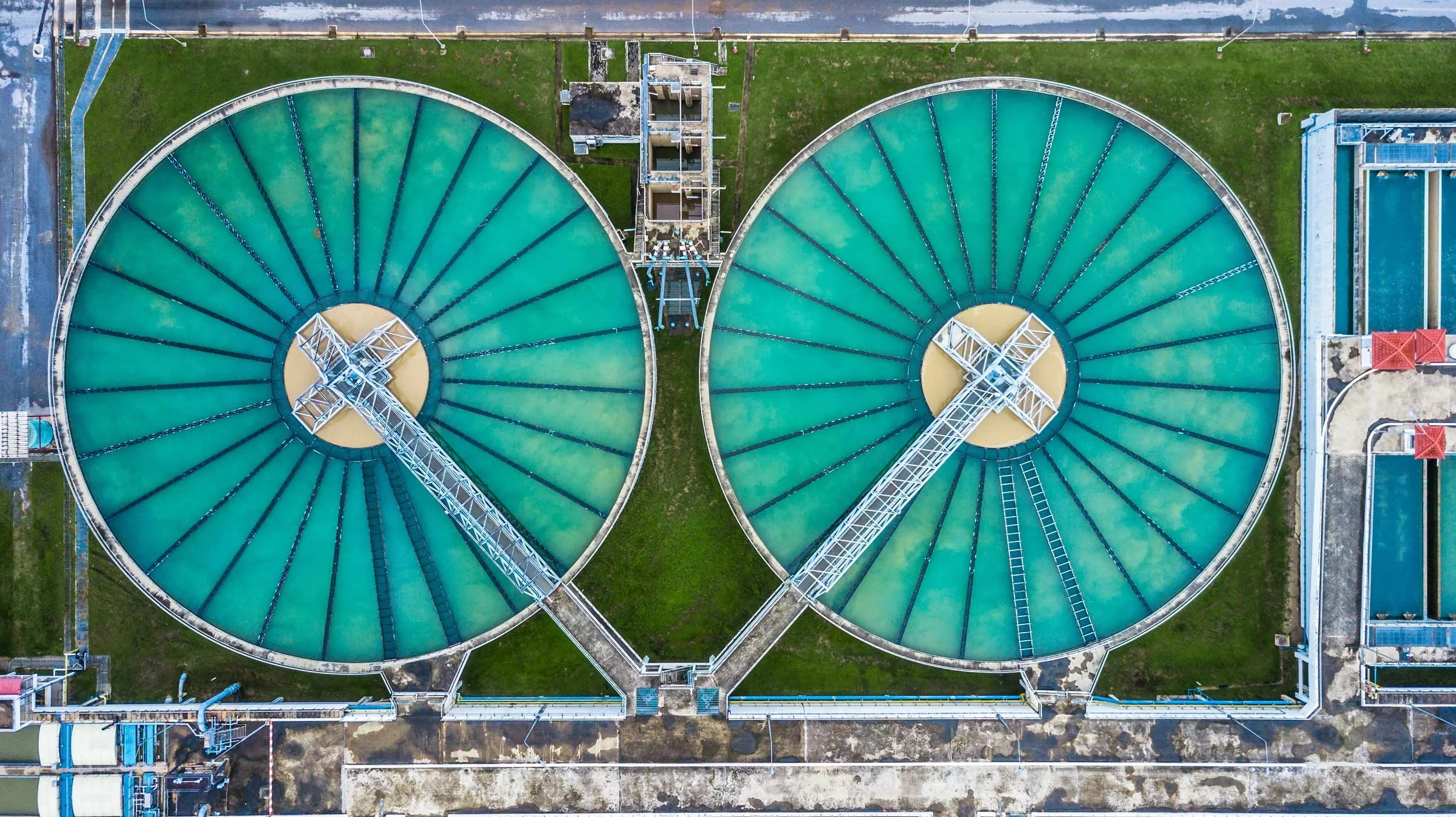Aerial view of two large circular water treatment tanks with greenish water, connected by a walkway, surrounded by industrial structures and green grass.