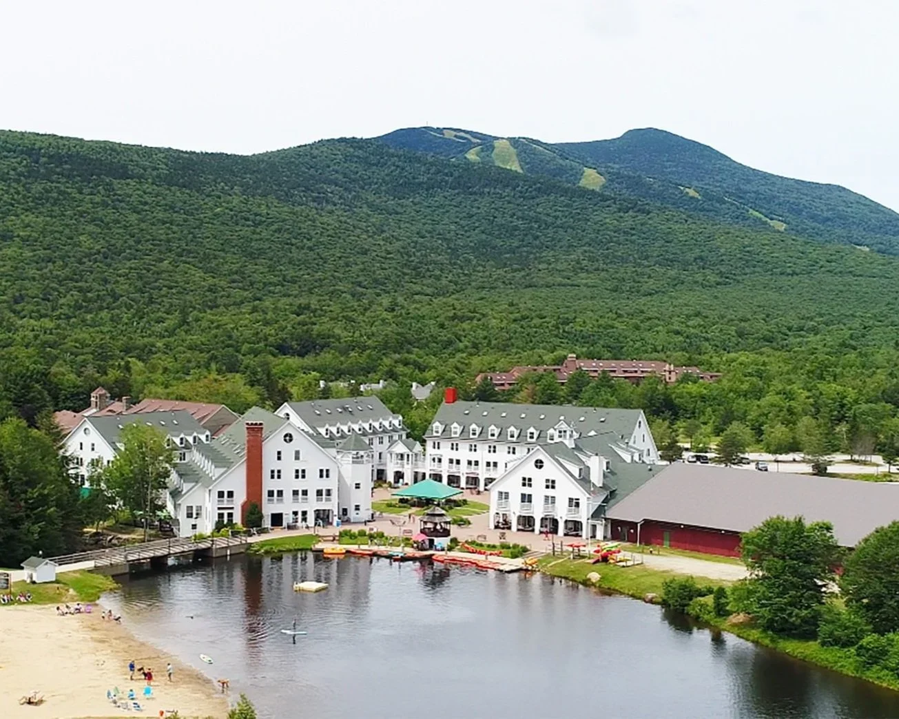 Resort near a lake with white buildings, green mountain backdrop, and a sandy beach with people, pod-style floats on the water, and paddleboards