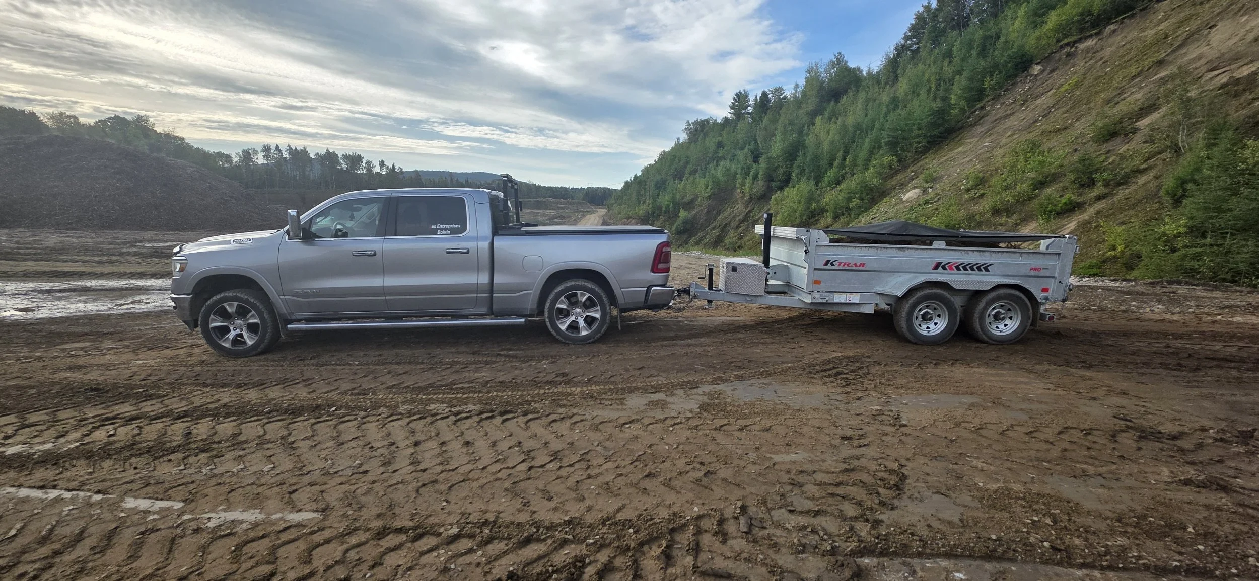 Une camionnette grise attachée à une remorque en terrain meuble avec une forêt en arrière-plan.