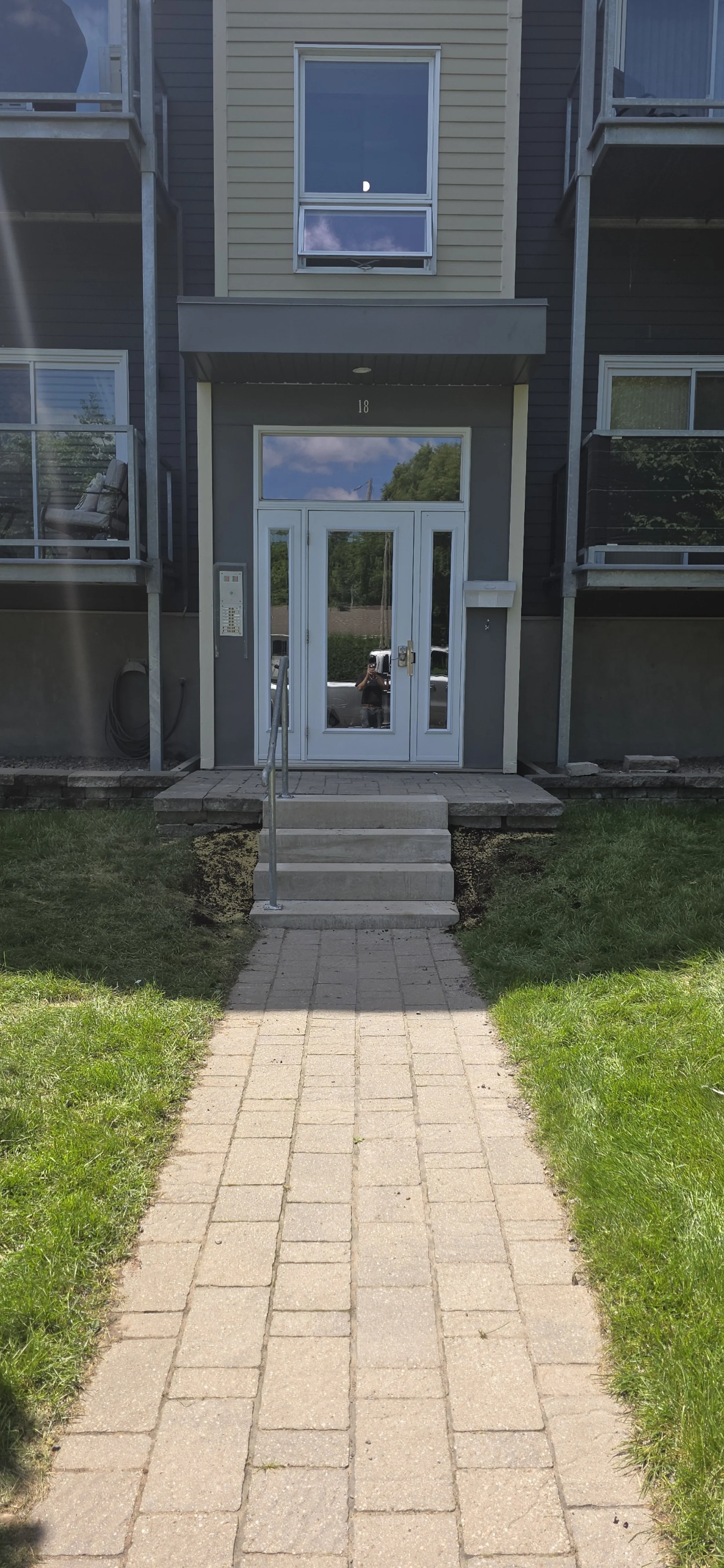 Vue de la façade d'un immeuble résidentiel avec une allée en pavés menant à l'entrée, un escalier avec rampe en métal, porte d'entrée en verre et fenêtres, un balcon sur chaque côté et la lune visible dans le ciel.