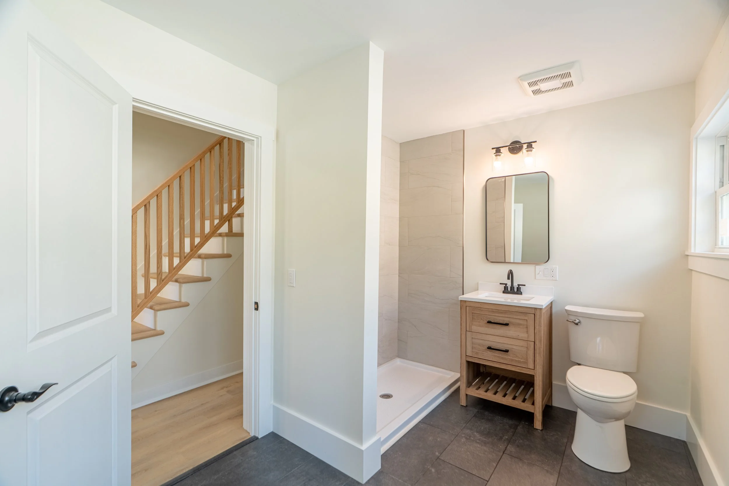 Small modern bathroom with white tiled walls, a wall-mounted small sink with a gold faucet, a mirror above, a toilet to the left, a shower area with a gold shower rod, a wall niche with hexagon tiles and bottles, a wall outlet, and a gold towel hook.