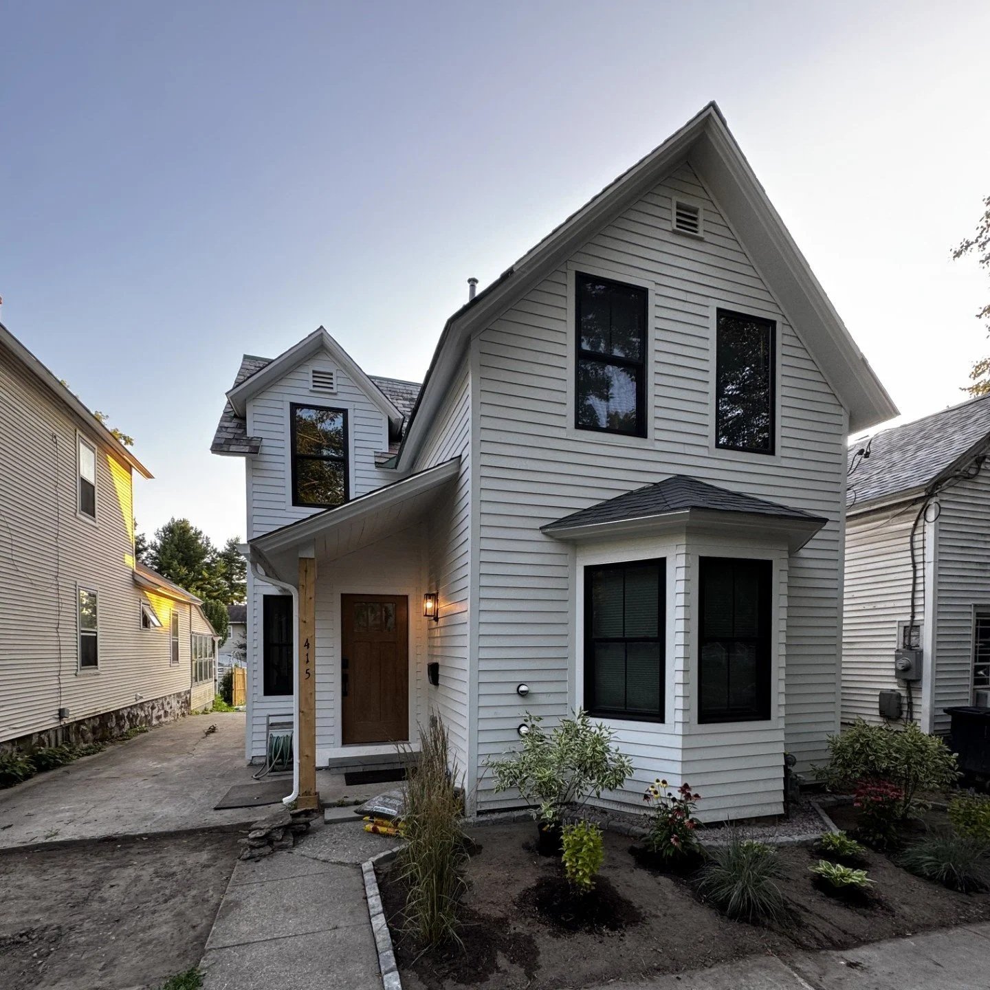 A newly constructed house with blue vinyl siding, four large white-framed windows, white lattice skirting, and a wooden deck stairs on the right side, surrounded by a dirt yard and trees in the background.