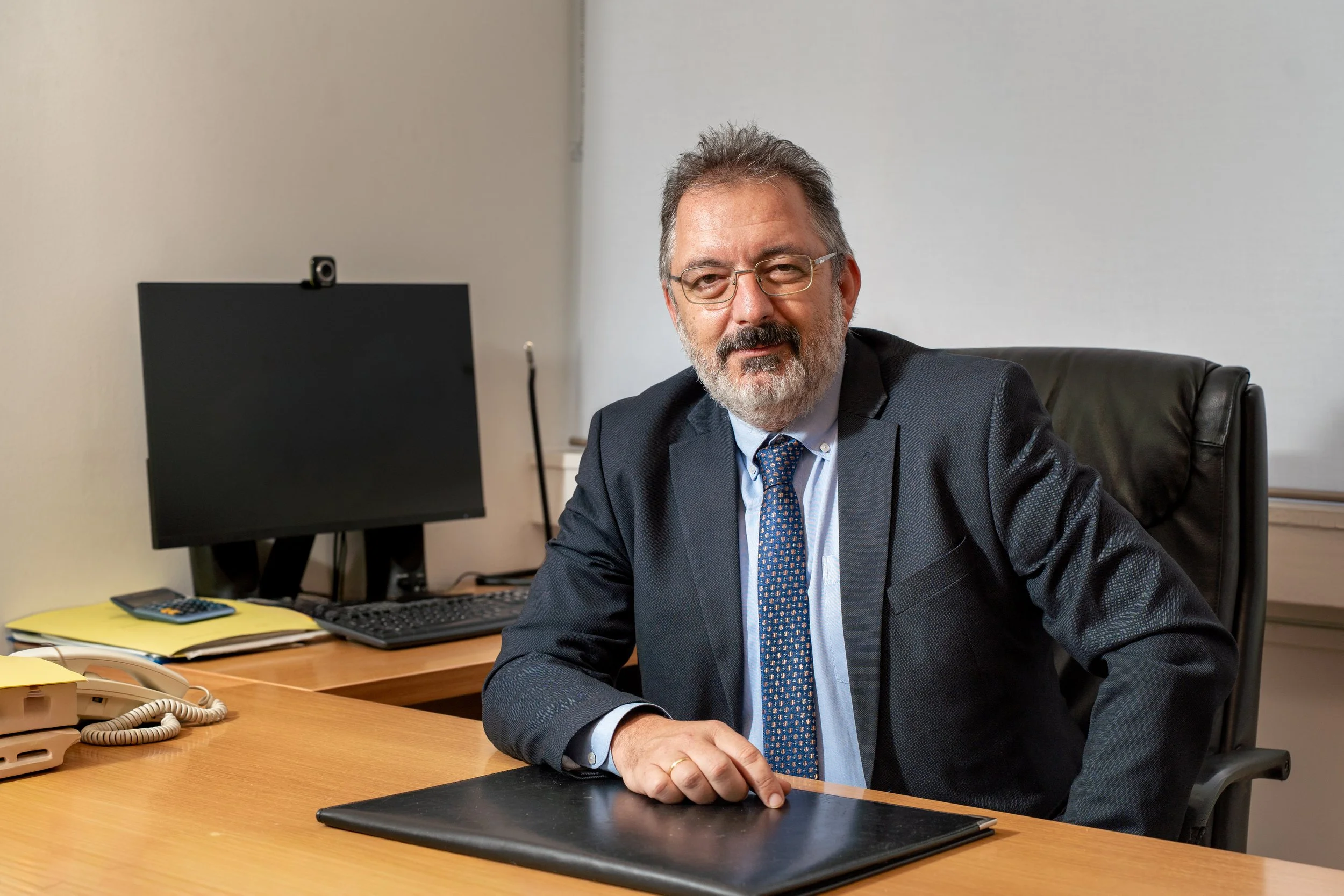 A middle-aged man with glasses, a beard, and gray hair, wearing a dark suit, light blue shirt, and patterned tie, sits at a desk in an office with a computer monitor, keyboard, and office phone.
