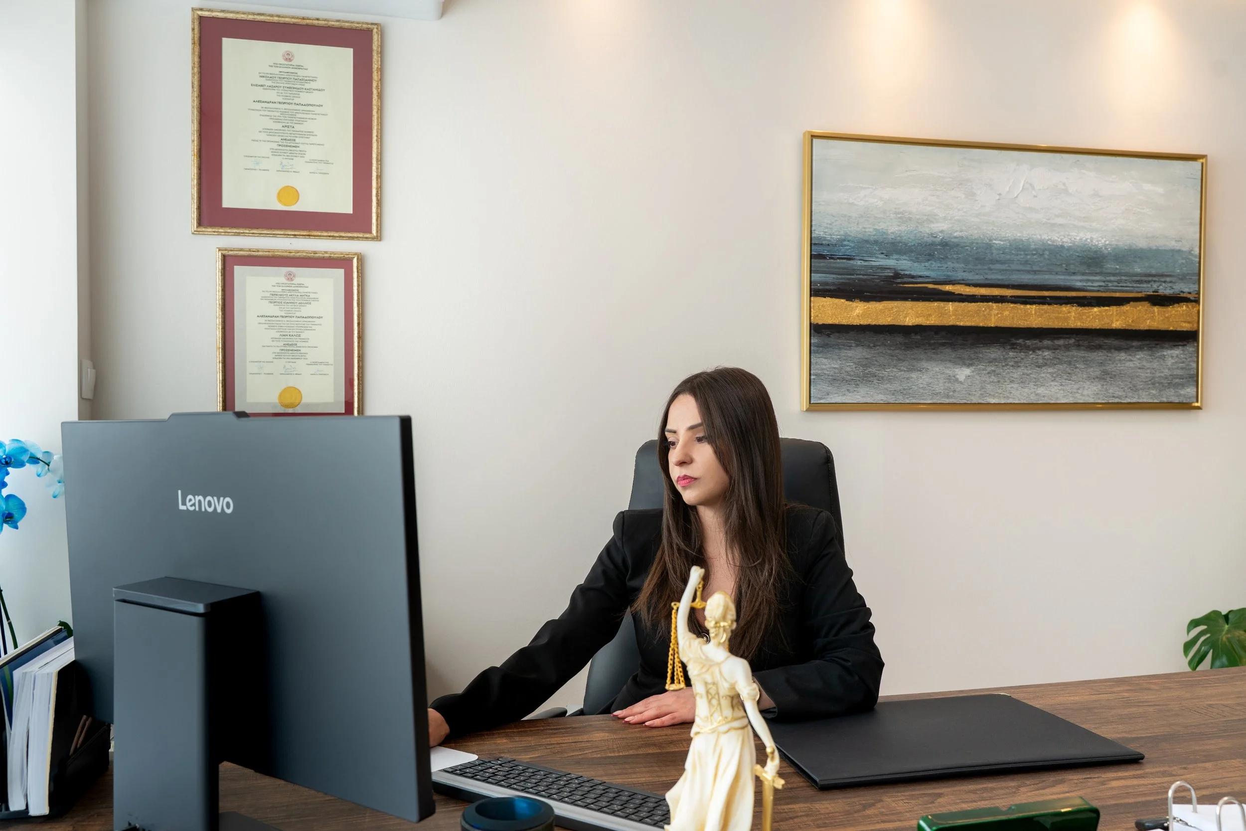 A woman sitting at a desk in an office, working on a computer with a Lenovo monitor. The desk has a statue, a pen holder, and a black folder. Behind her on the wall are framed certificates and an abstract painting.