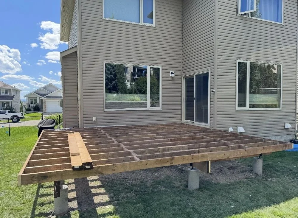 Unfinished wooden deck framework attached to the back of a beige house with vinyl siding, in a suburban neighborhood.