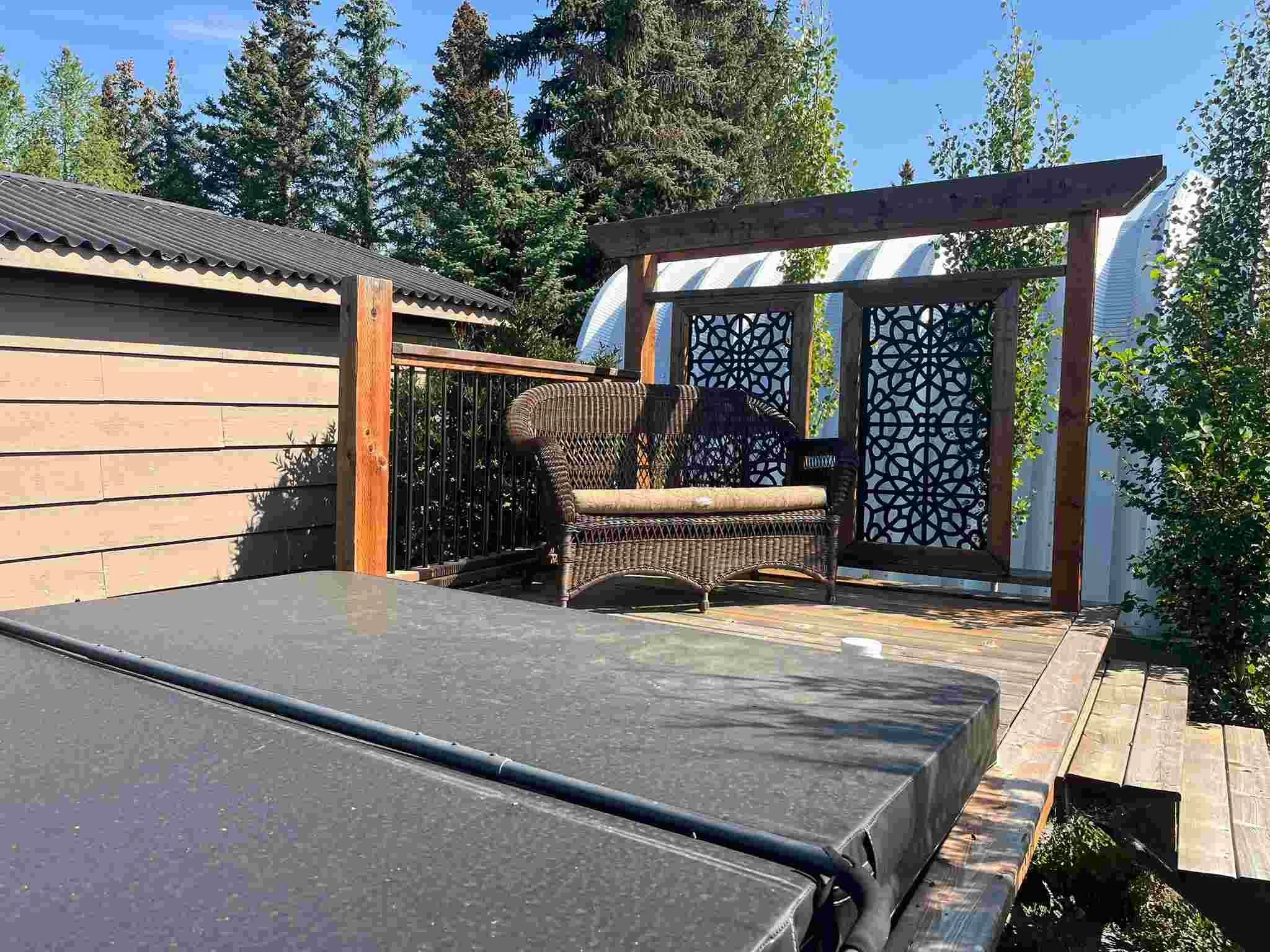 A small wooden deck with a wicker loveseat and a table with a black cover. The deck has decorative black panels, a wooden railing, and is surrounded by green trees under a blue sky.