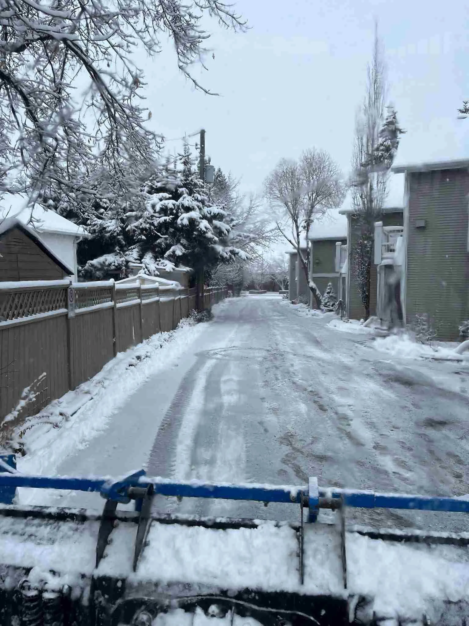 View from a snowplow on a residential street showing snow-covered road, trees, houses, and fences, with overcast sky.