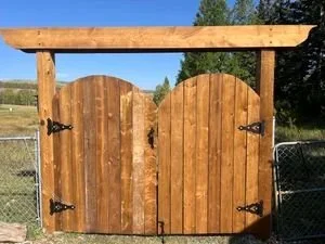 Wooden garden gate with curved top, decorated with black hinges, set between two wooden posts in an outdoor setting with trees and a chain-link fence in the background.