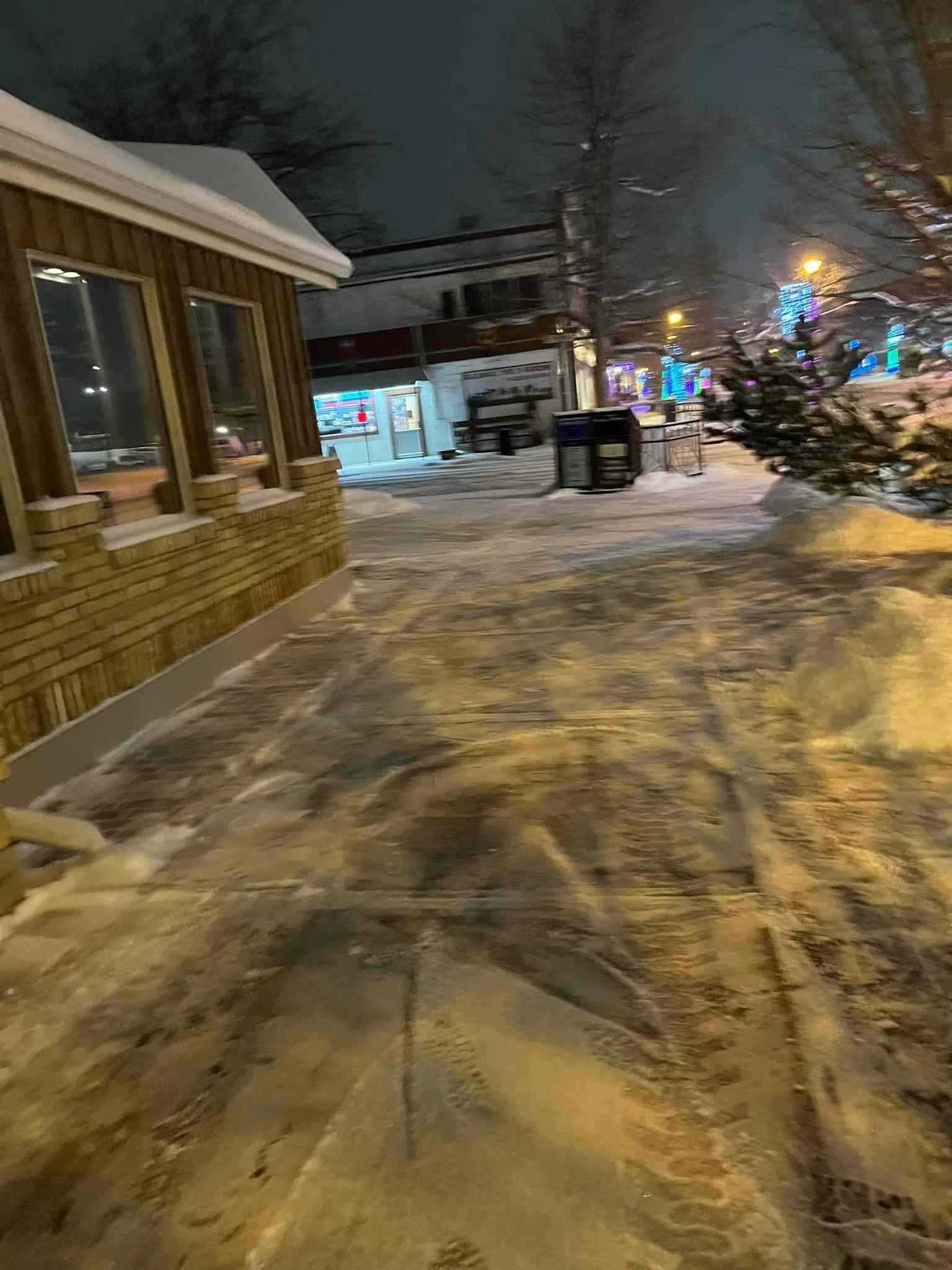 Snowy sidewalk near a brick building with windows, trees, and colorful holiday lights at night.