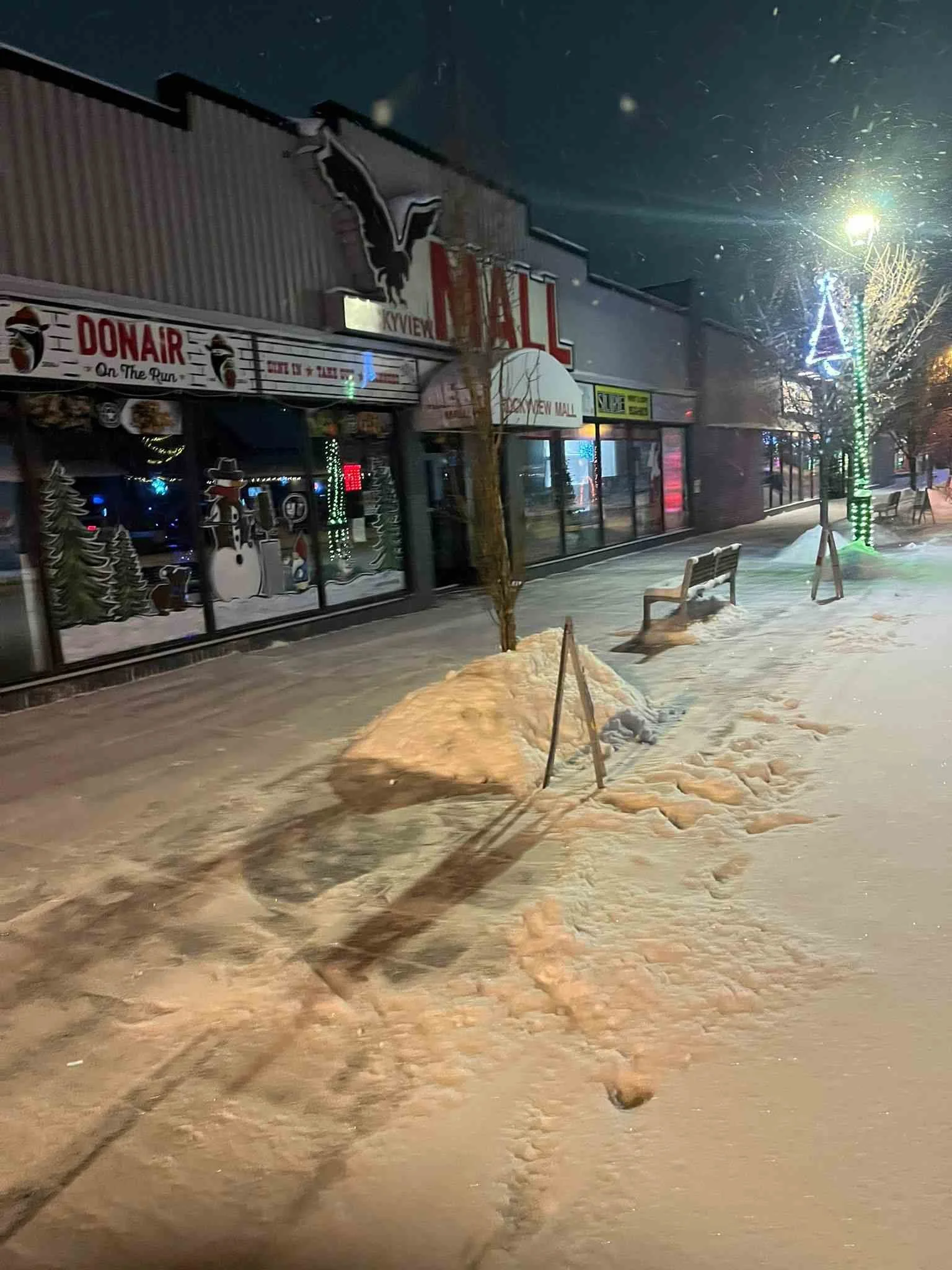 Snow-covered sidewalk outside a mall at night, with benches, trees decorated with holiday lights, and storefronts illuminated, including a sign for 'KXVIEW Mall' and an ice skating rink decoration visible through the window.