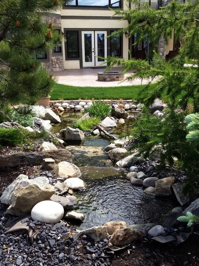 Custom backyard landscaping with a natural stone water feature, flowing stream, pond area, river rock, and surrounding greenery in front of a patio
