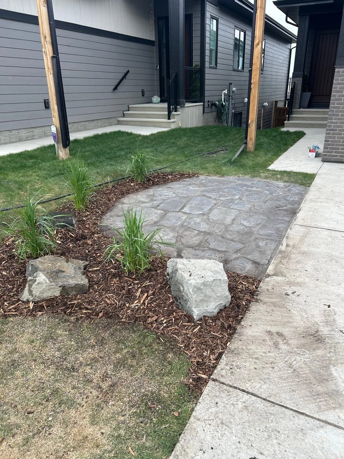 A front yard landscaping project showcasing a newly installed gray flagstone patio extension next to a concrete walkway, accented by dark mulch beds, natural stone boulders, and ornamental grasses