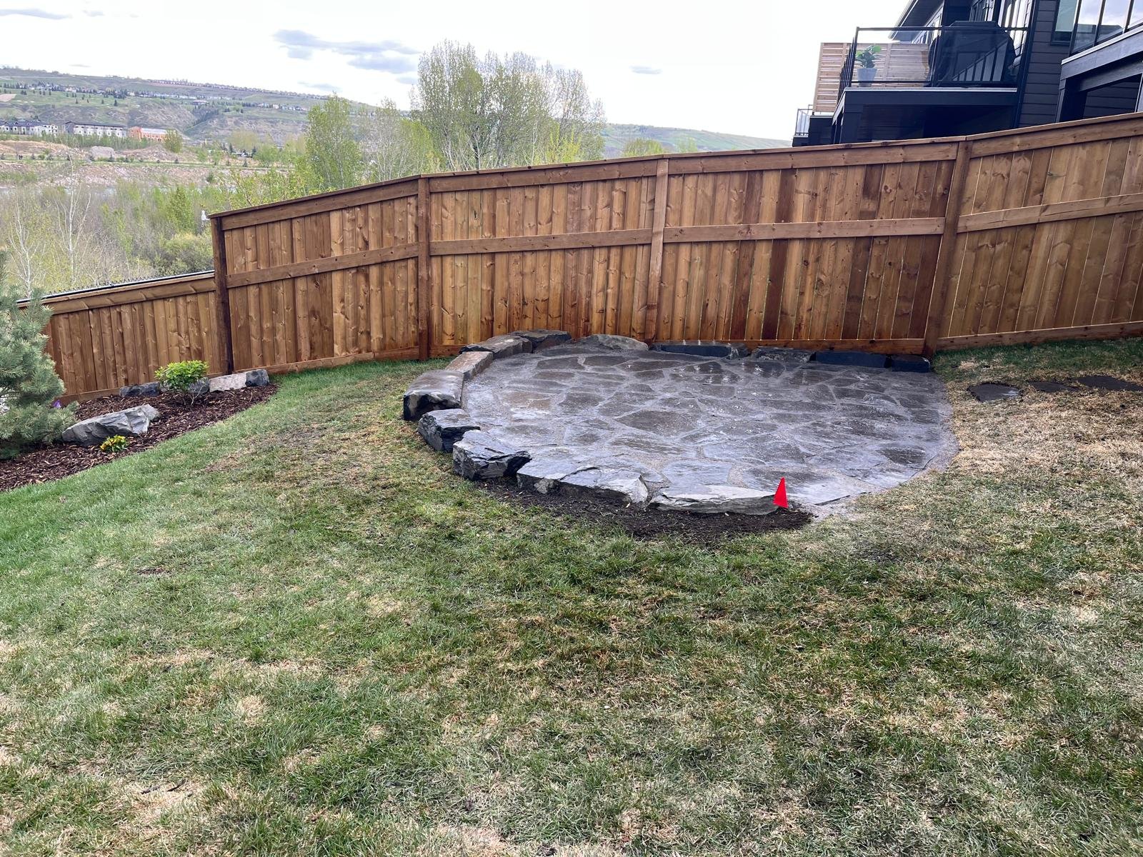 A completed backyard hardscaping project featuring a level circular flagstone patio built into a green grassy slope, supported by a heavy natural stone boulder retaining edge next to a tall wooden fence