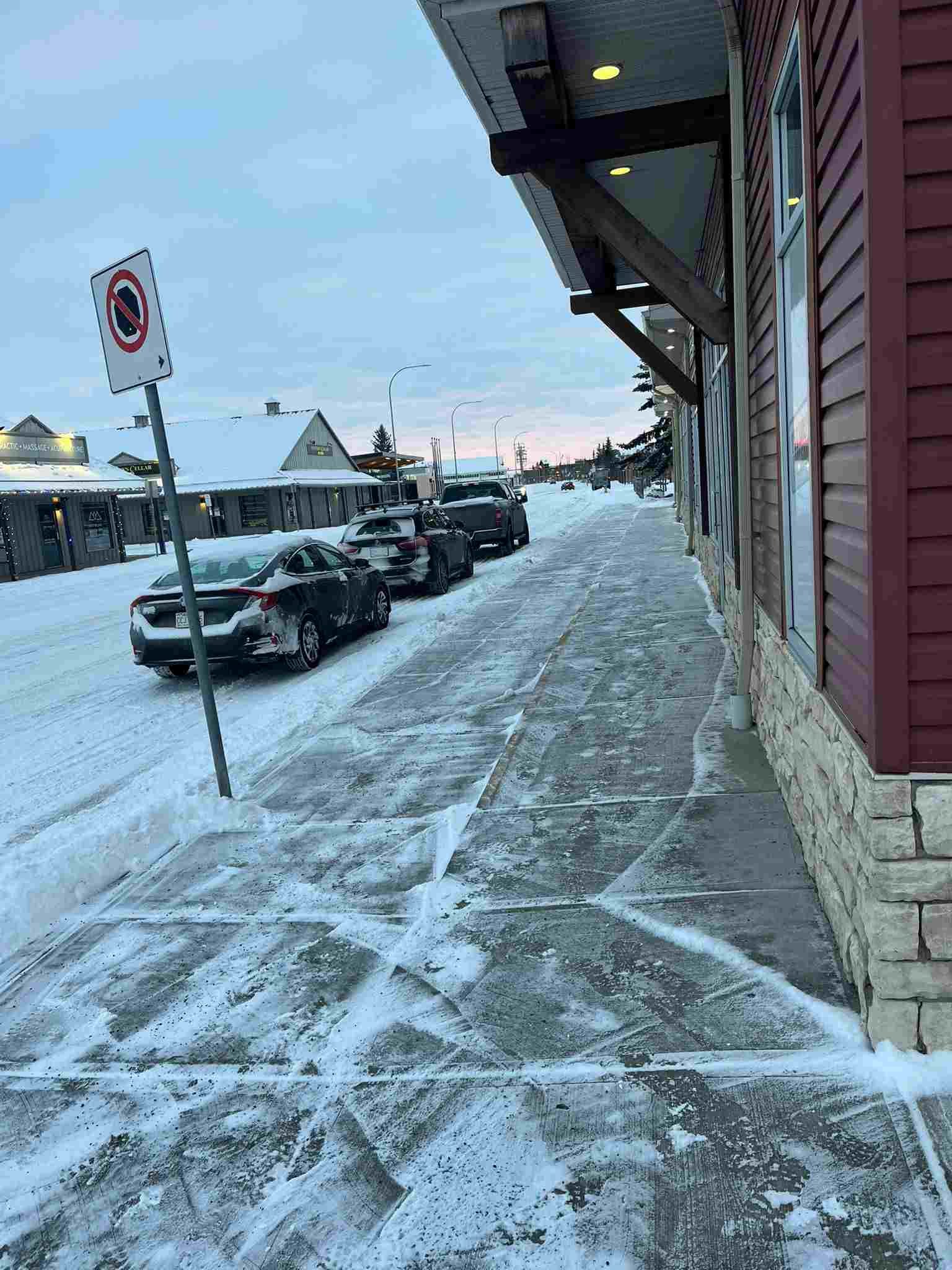Snow-covered sidewalk and parked cars along a street in a small town or neighborhood, with a building on the right side and a no parking sign in the foreground.