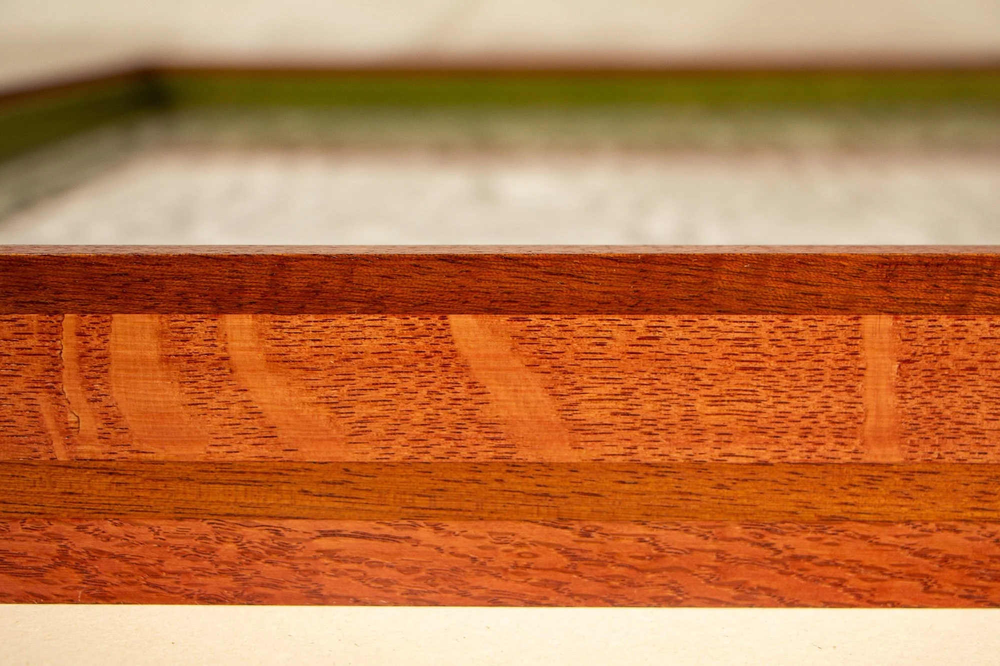 Resonant Syntax 003 frame: close-up of the mitered outer corner of the layered mahogany-and-oak frame, with red and granada-toned bands, visible grain, and the green inner layer softly receding in the background