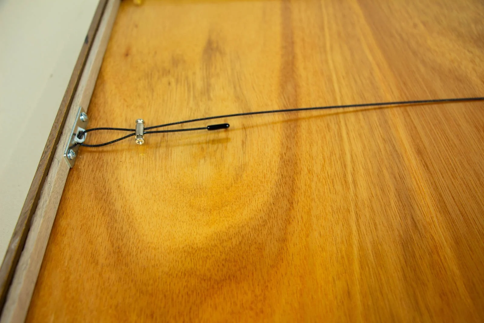 Synaptic Mythos 003 frame: hanging hardware close-up, steel plate with twin screws; black-coated wire looped and secured by a nickel compression barrel (with end sleeve); warm honey backboard grain