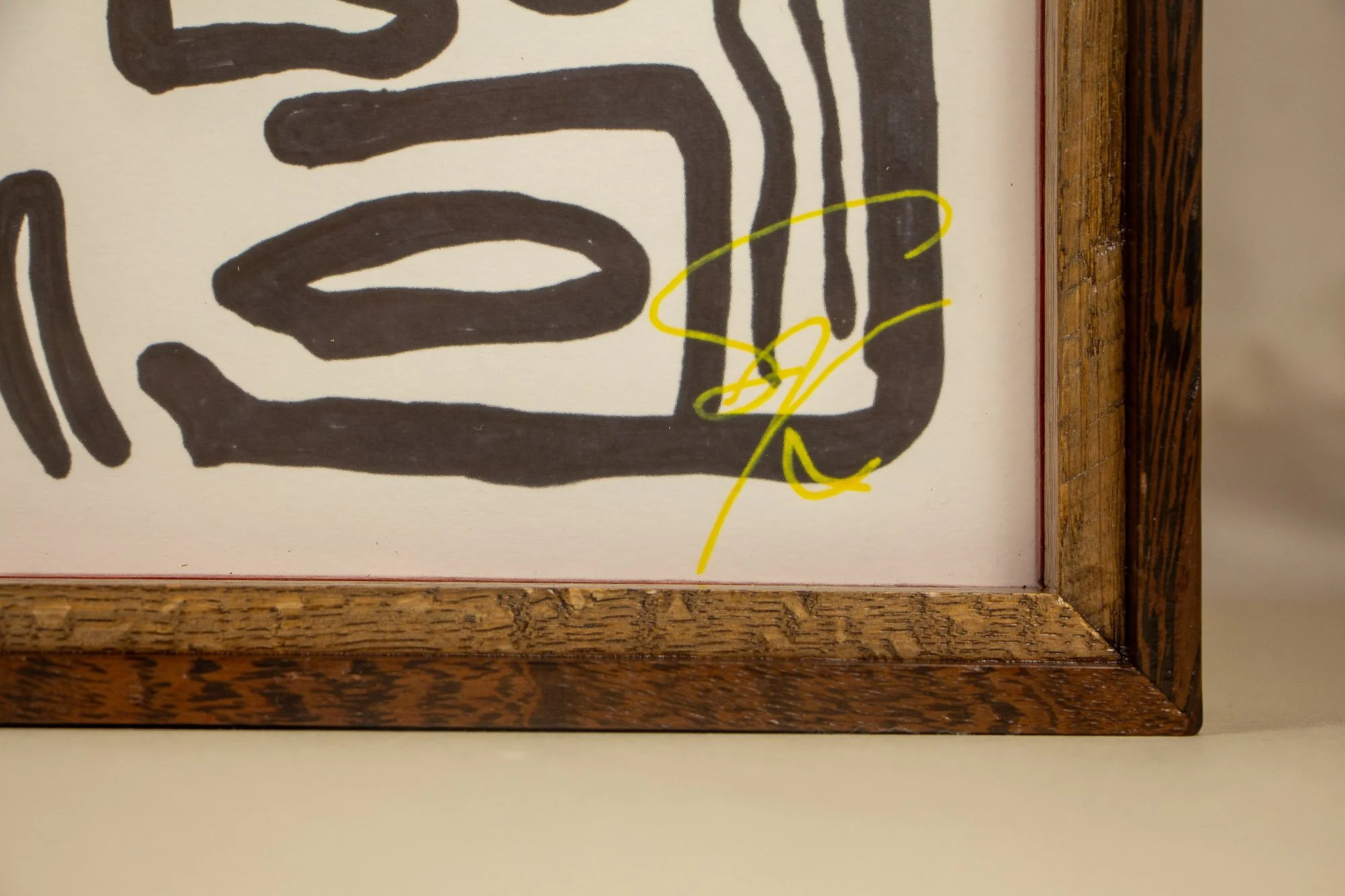 Elusive Cocoon 001 frame: lower-right front corner with the artist’s yellow signature behind glass, framed by the light ash-toned inner rail and red liner.