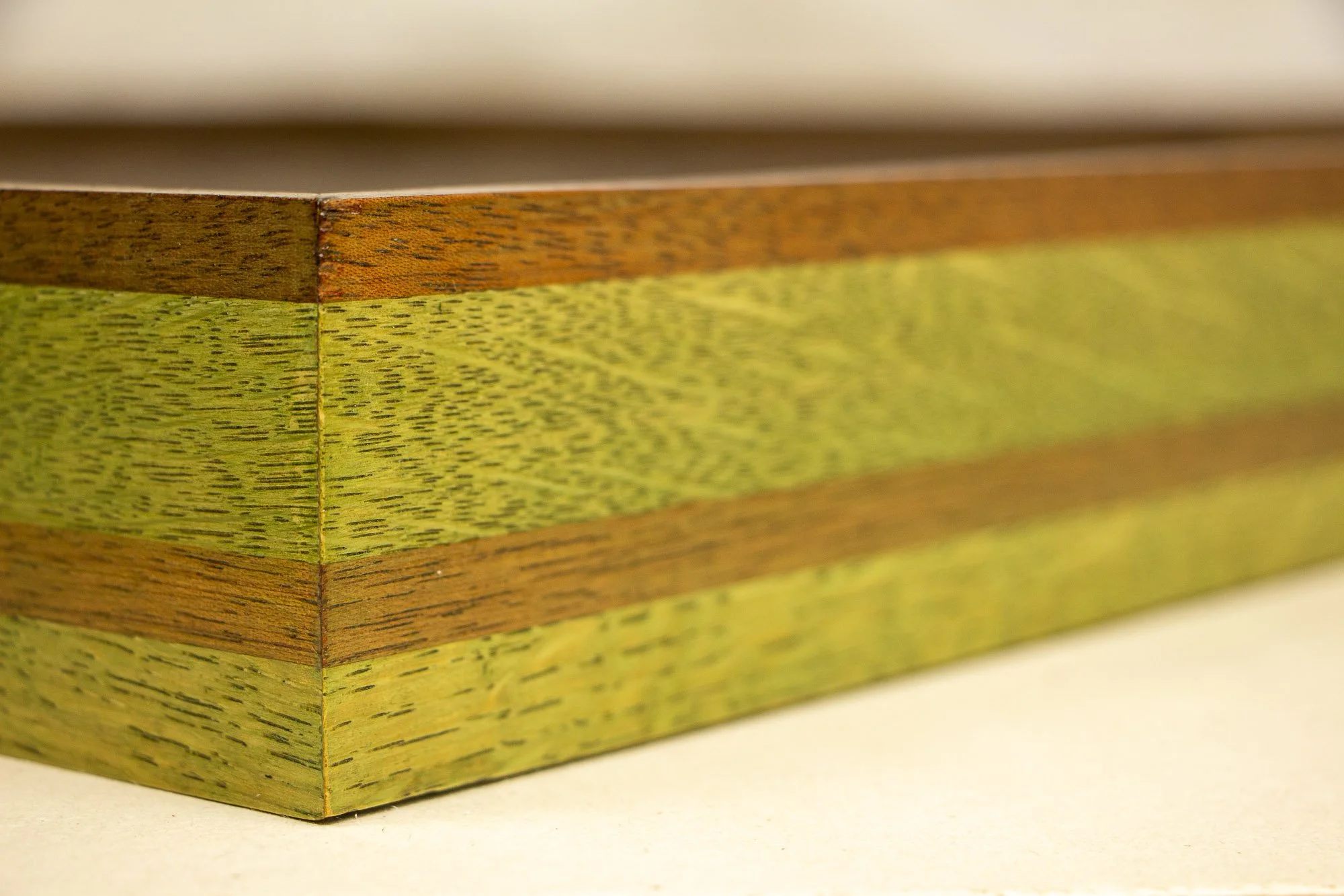 Resonant Syntax 004 frame: close-up of the mitered corner of the layered mahogany-and-oak frame, with the green, granada, and red sequence visible through contrasting stained bands and aligned grain