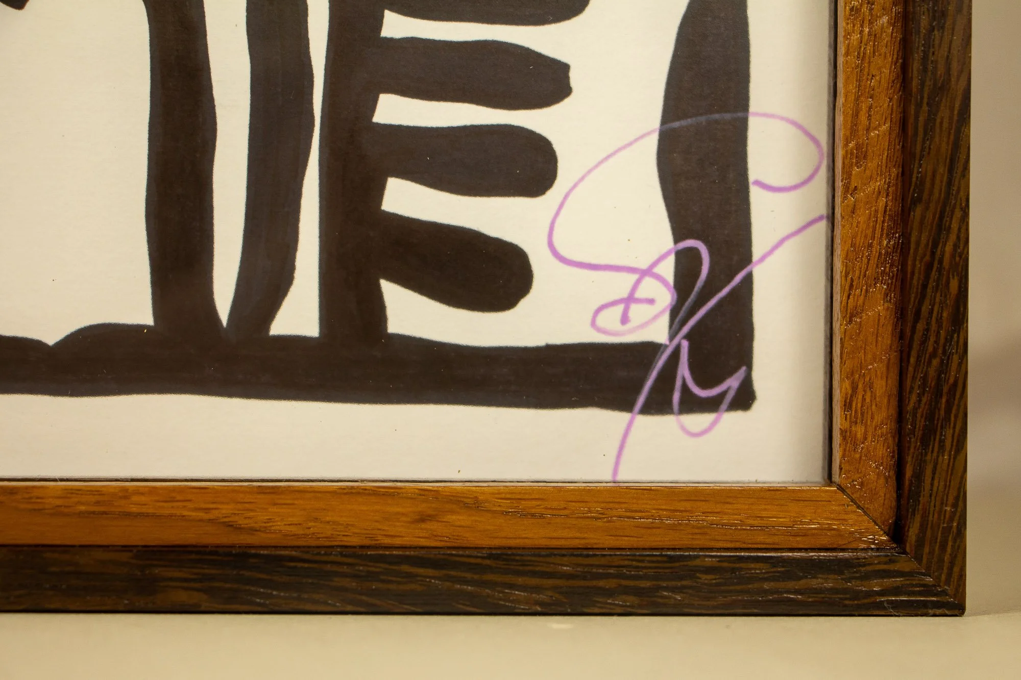 Elusive Cocoon 002 detail and frame: close-up of the lower signed corner with purple signature, bold black linework on warm white paper, and the layered wooden frame surrounding the work.