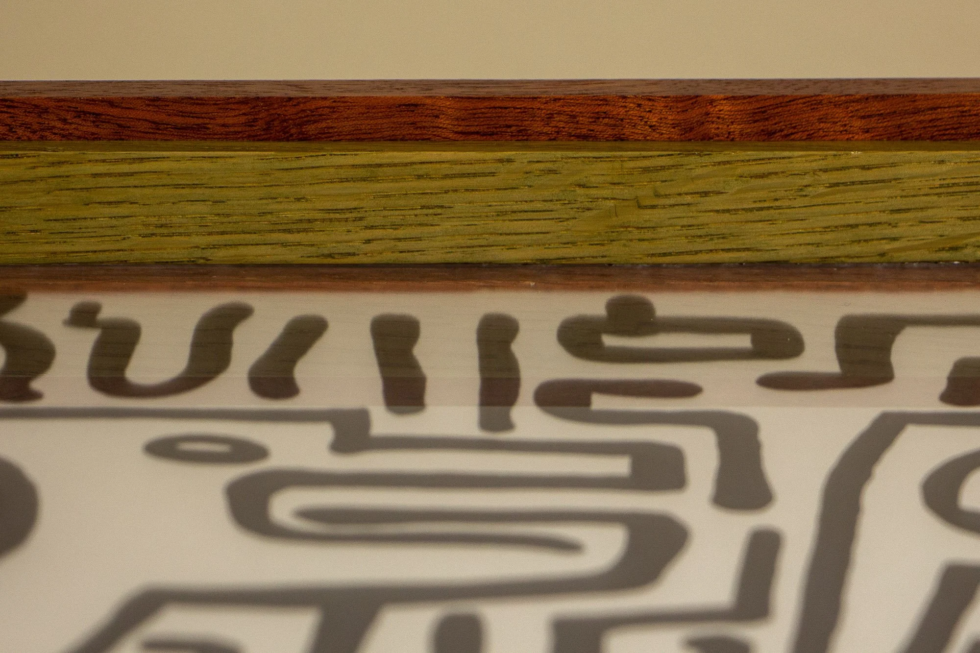 Resonant Syntax 001 frame: upper edge detail with mahogany outer frame, colored oak spacer elements, and black ink linework visible beneath the glass