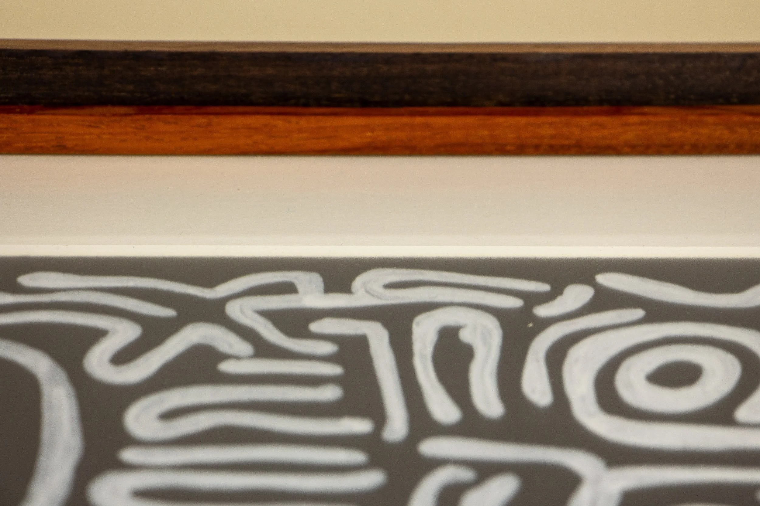 Mêlée of Discovery 001 frame: shallow-focus close-up of the inner frame edge and glazing, where the warm mahogany lip meets the off-white surround and the artwork dissolves softly beneath it.