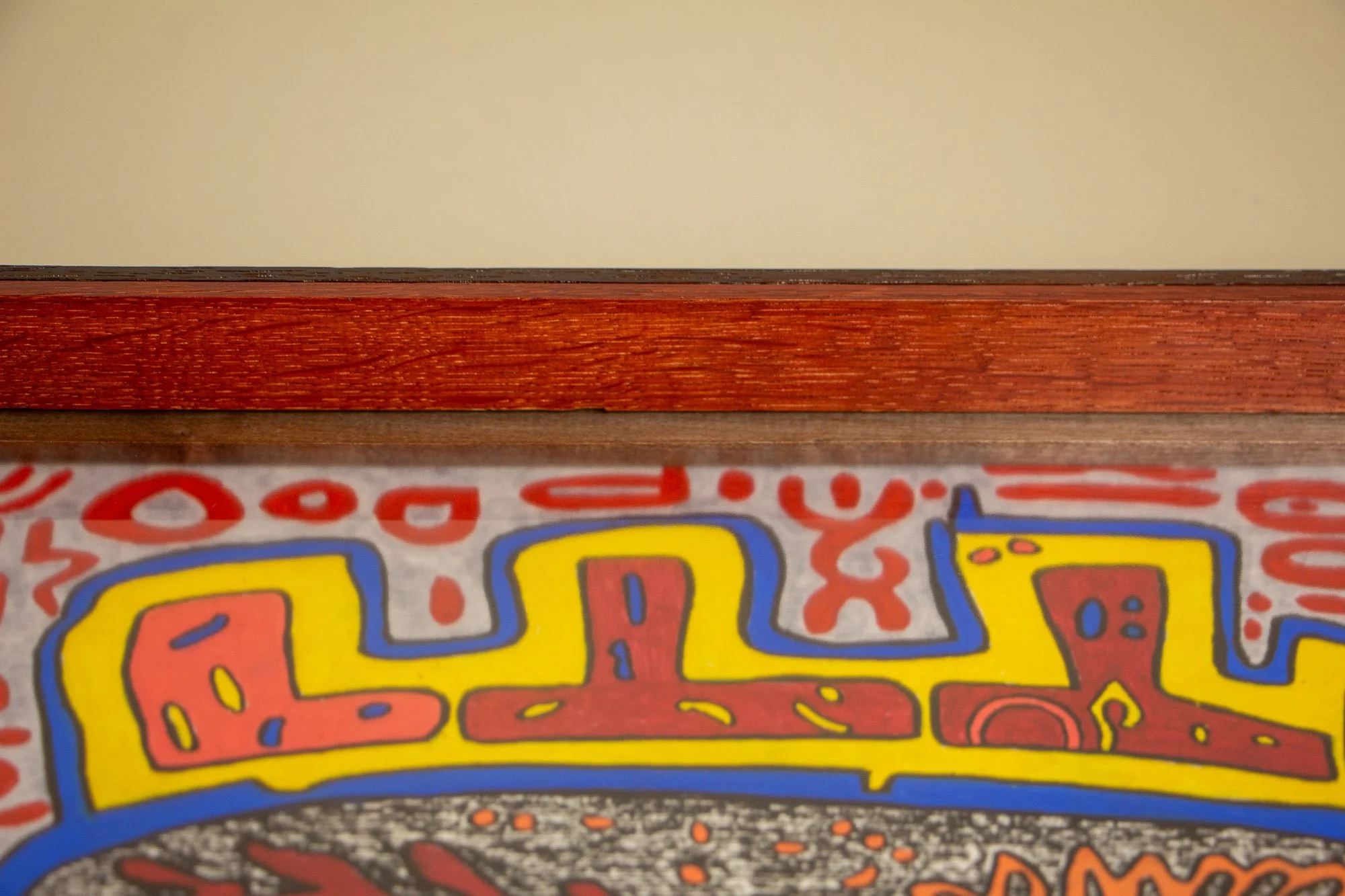 Close-up of a colorful graffiti-style painting on a wall, with abstract shapes and bright colors including yellow, red, blue, and black, under a wooden shelf or ledge.