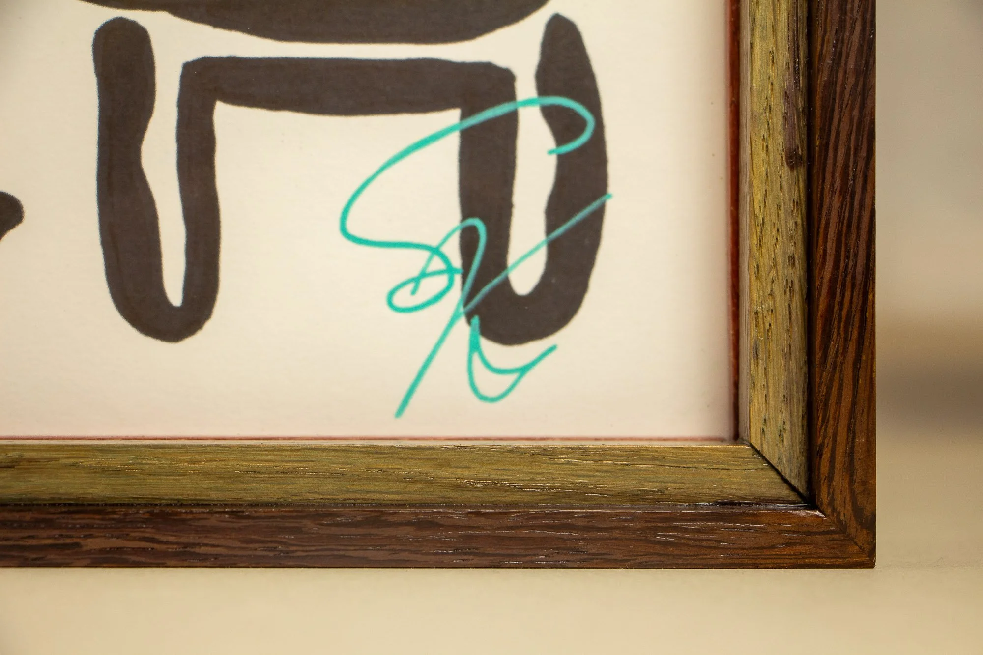 Elusive Cocoon 003 detail and frame: signed lower corner with turquoise signature on warm white paper, set inside a layered wengé-and-oak frame with mint-toned inner band and dark outer rail.