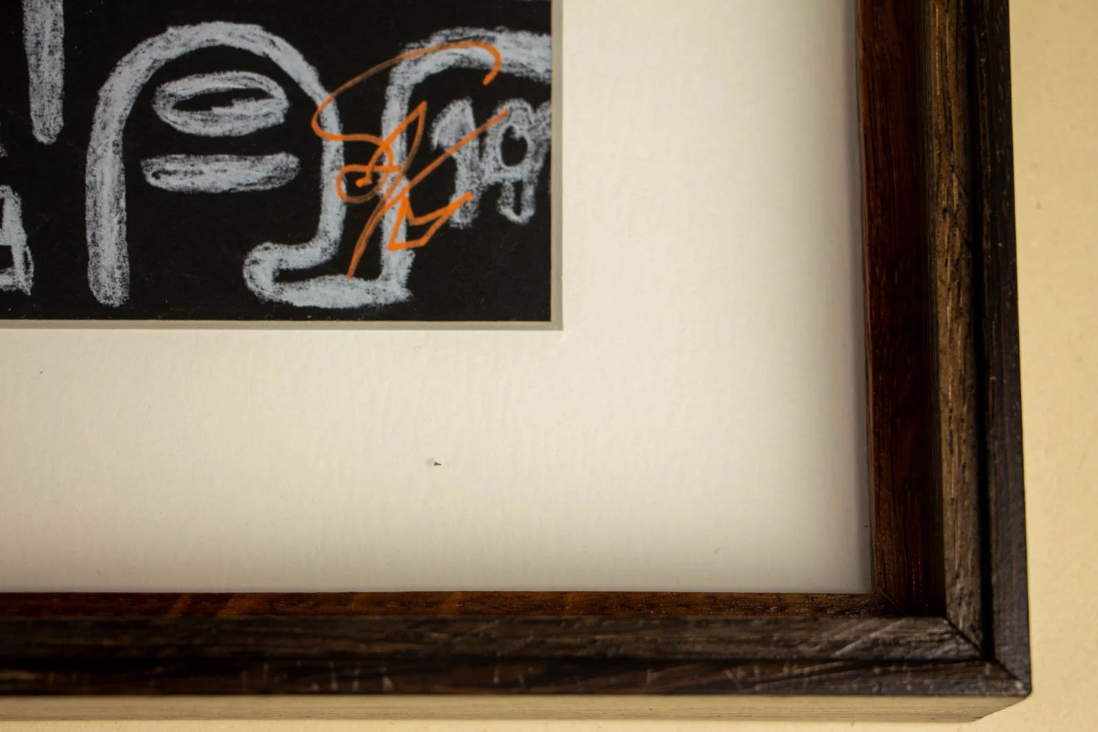 Close-up of the bottom right corner of a framed chalkboard sign with white chalk writing and a signature, hanging on a beige wall.