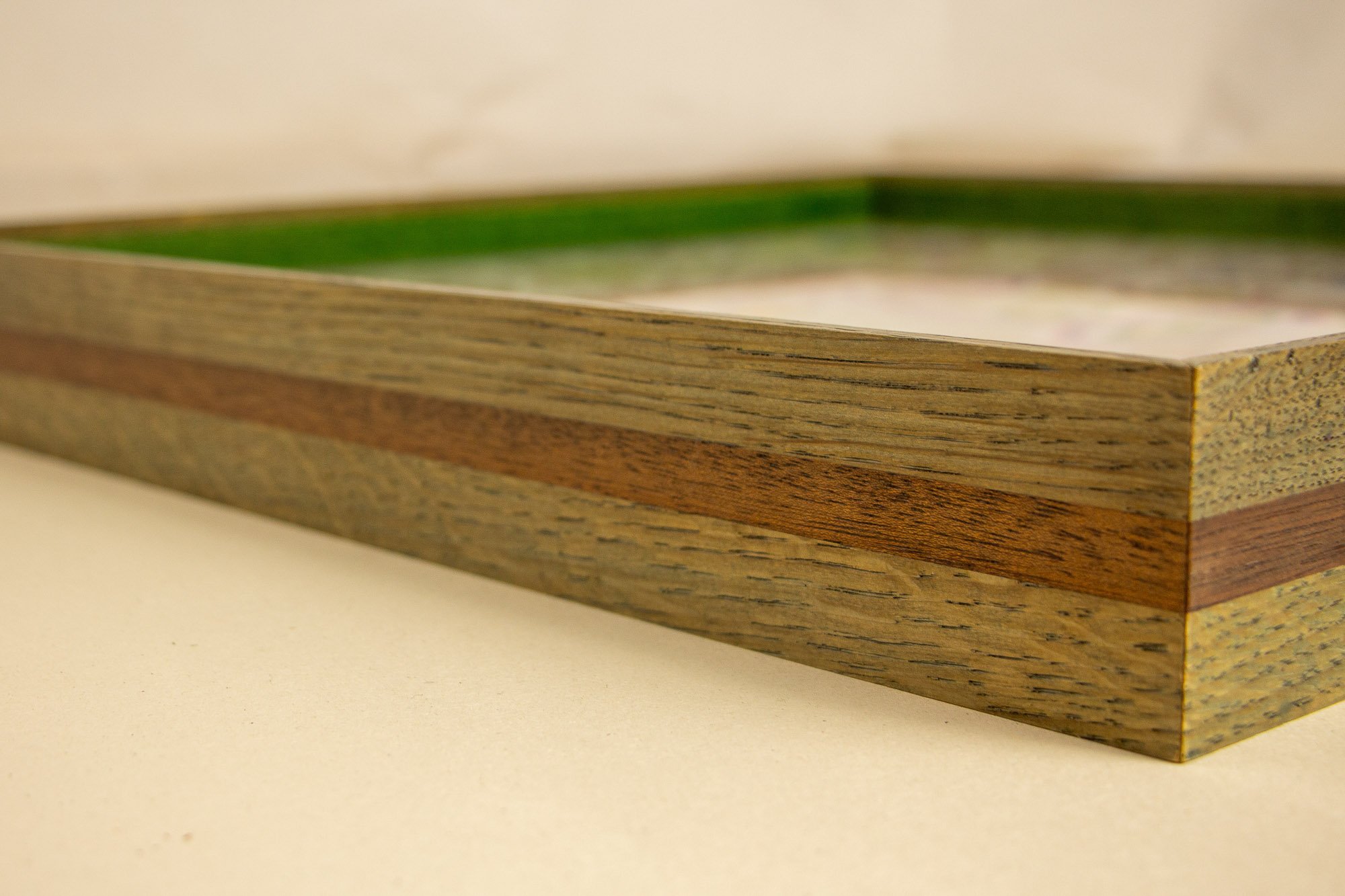 Eclipsing Gravitas 002 frame: angled side view of the shallow oak-and-mahogany frame profile, with olive-green toned oak faces, a warm central mahogany band, visible natural grain, and a bright green inner edge.