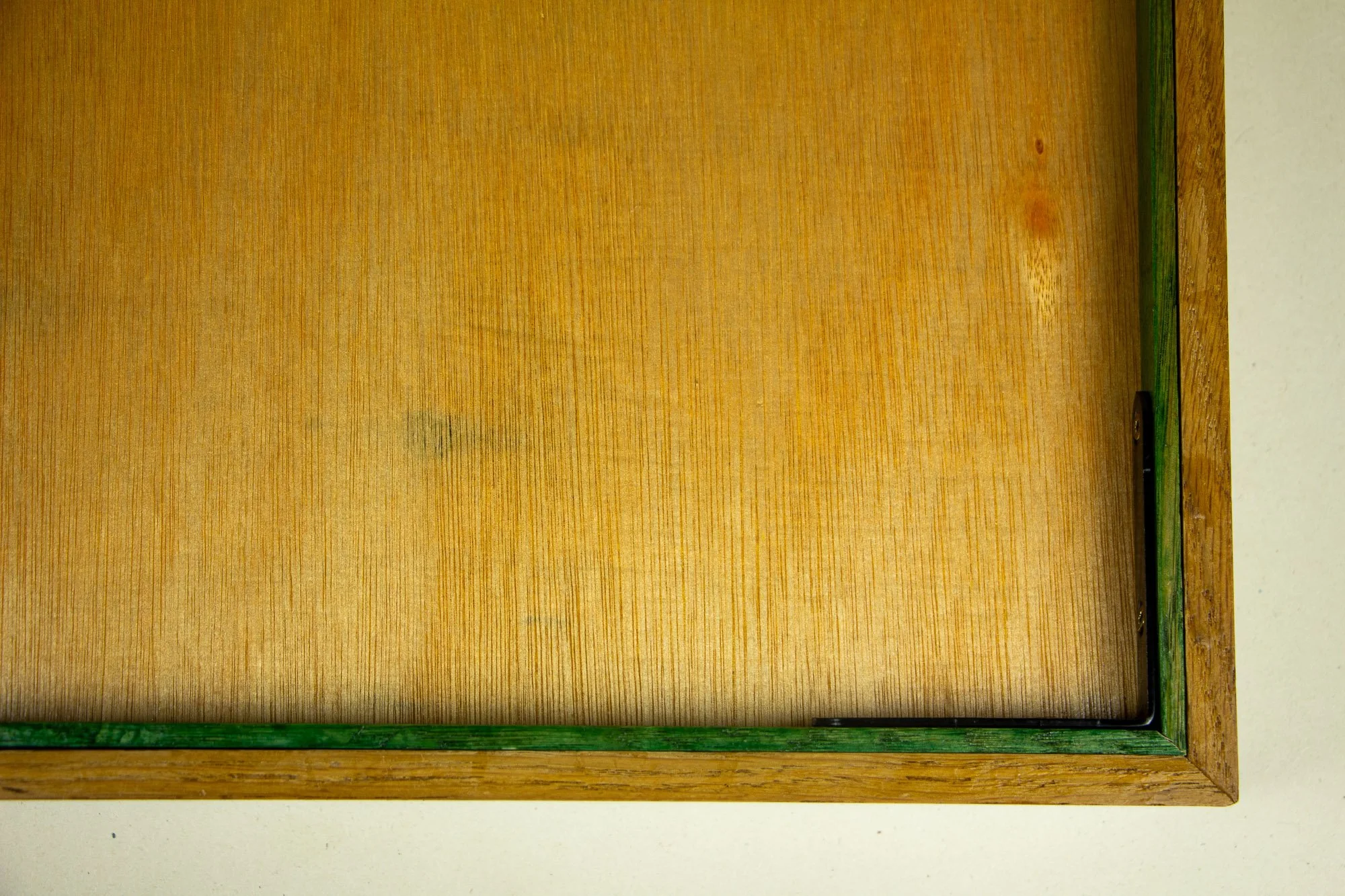 Elusive Cocoon 004 frame: overhead reverse-side corner detail showing the meranti plywood back panel, green-toned inner rim, outer oak edge, and black metal corner brace fixed into the frame construction.