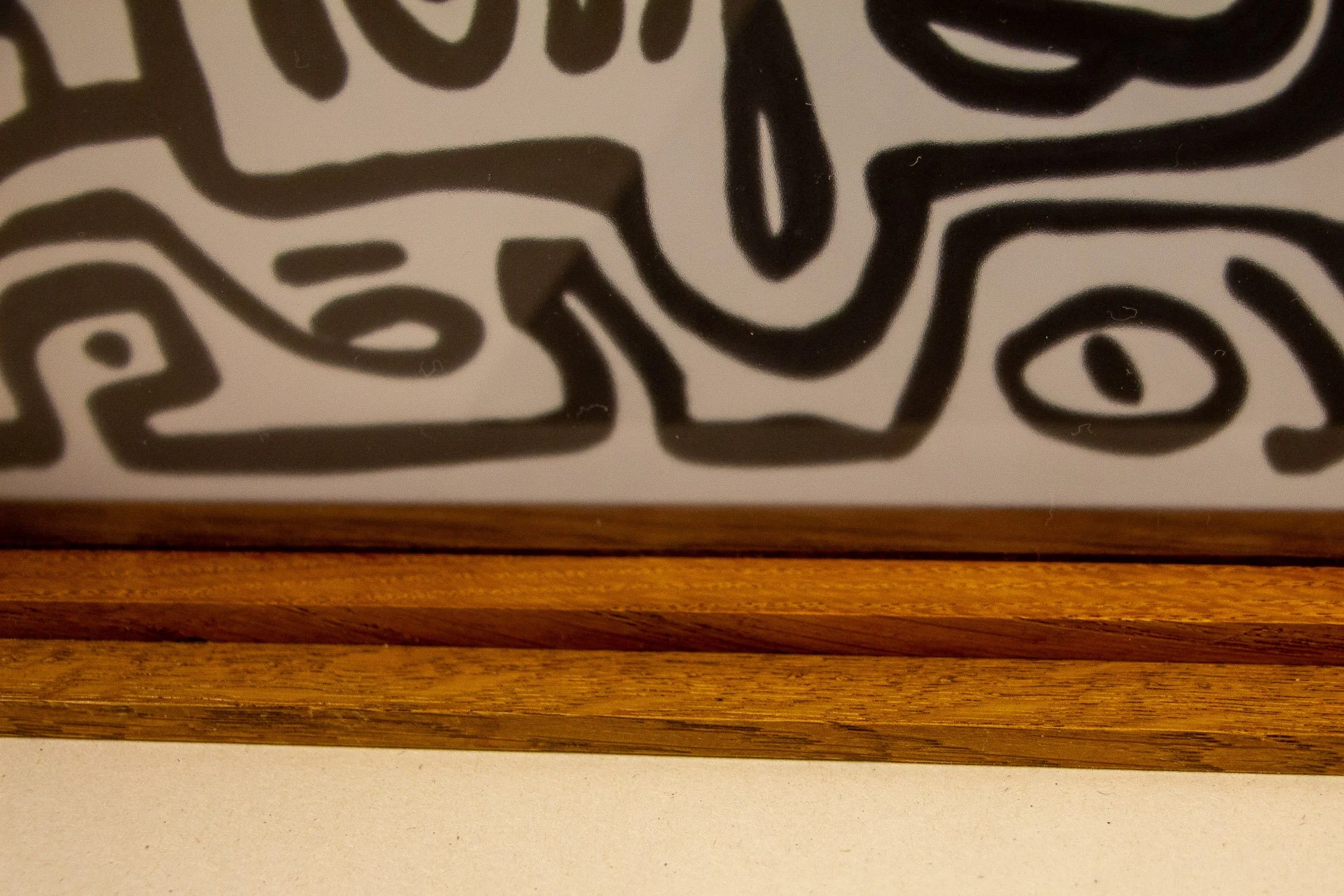 Veiled Alchemist 003 frame detail: front-facing close-up of the lower-left edge, showing the warm grain of the outer frame, the inner lip, and a cropped portion of the drawing with bold black linear forms.