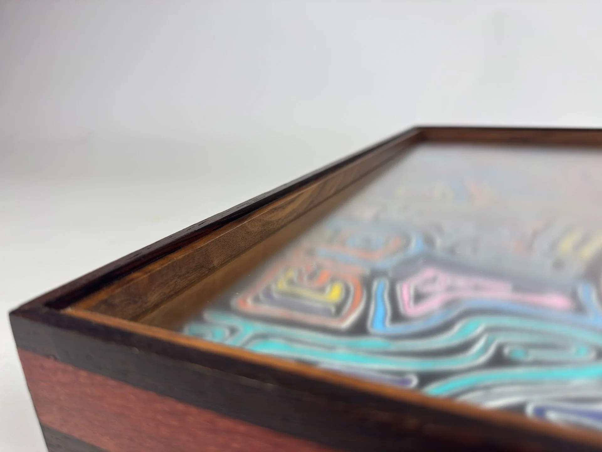 Corner detail of the custom frame, showing the mitered join in wengé, oak, and walnut, with pink, grey, turquoise, and orange line motifs from the artwork fading softly beneath the glazing.