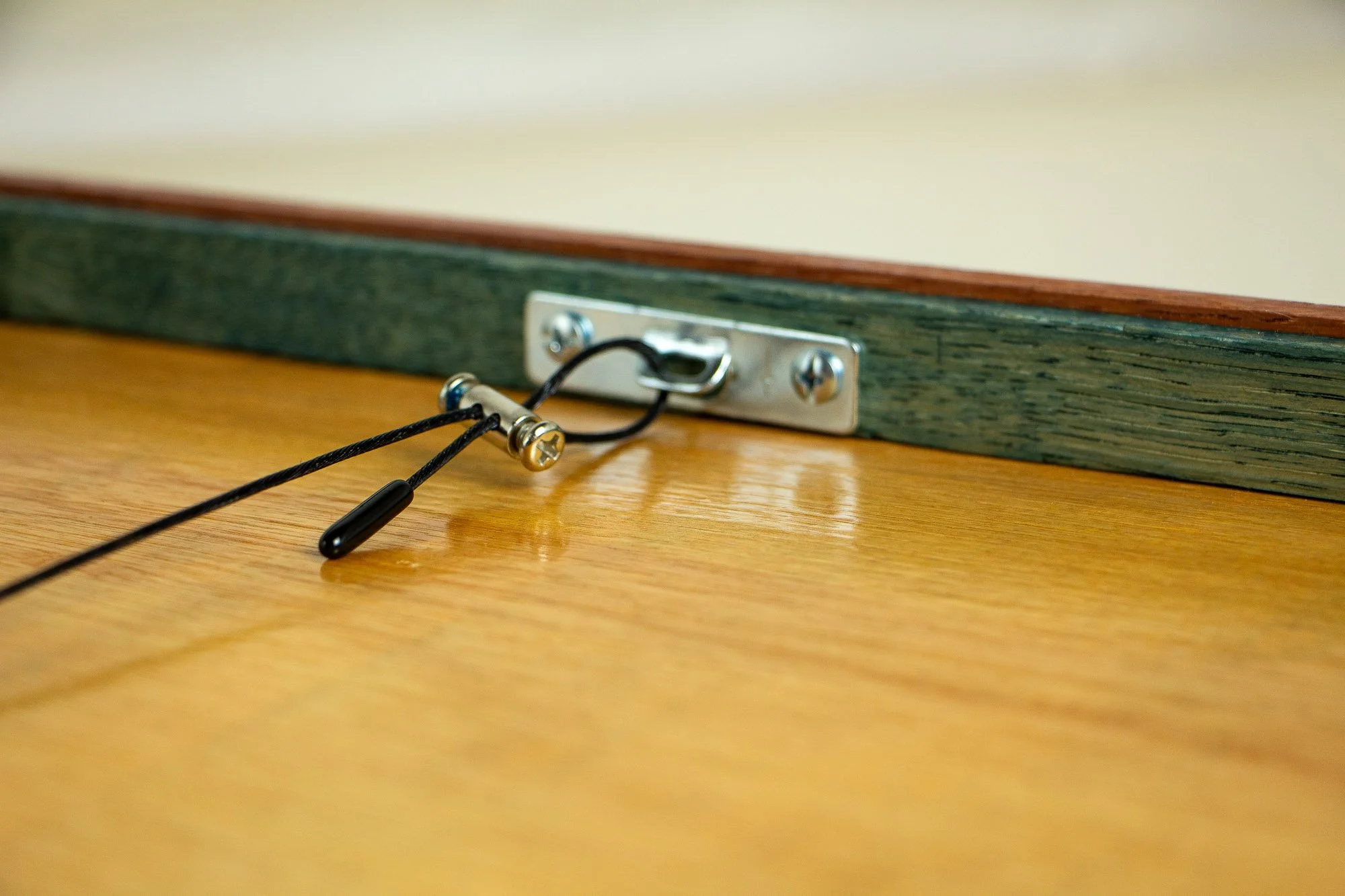 Unseen Signifiers 005 frame detail: angled reverse close-up of the hanging system, showing the metal fixing plate, black hanging cord, adjustable tension fastener, warm plywood back panel, and the olive-green inner support rim along the frame edge.