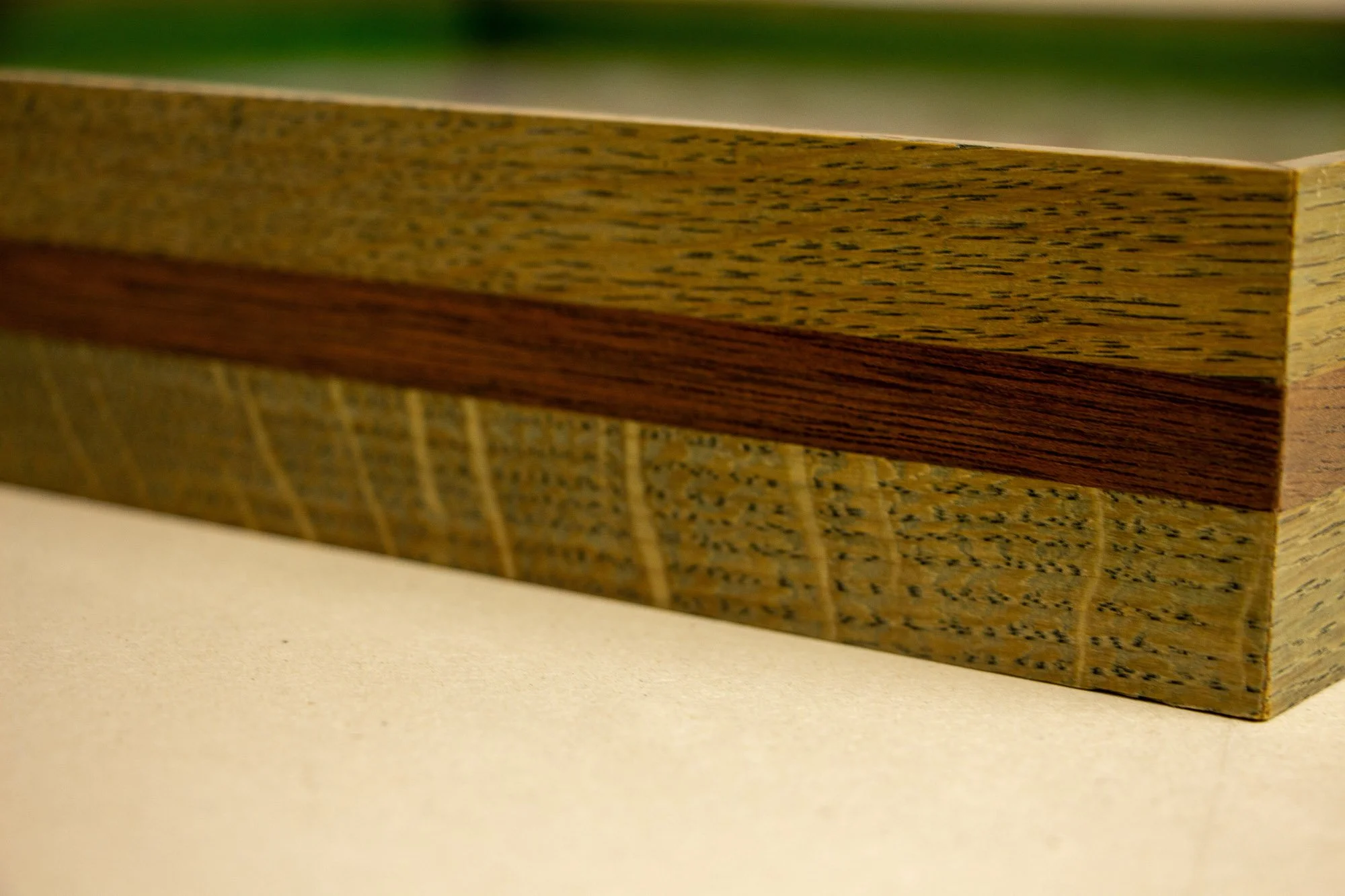 Eclipsing Gravitas 002 frame: frontal macro of the frame profile with olive-green toned oak and a warm mahogany band, showing the low-profile construction, visible grain variation, and the artwork softly blurred beneath glass.