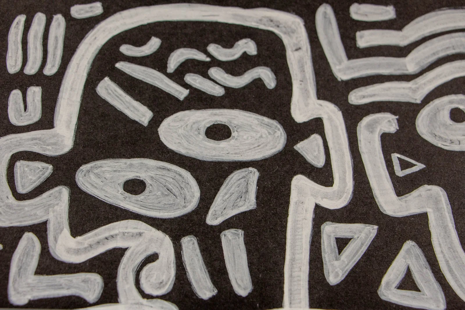 Metamorphic Entwining 002 detail: mask-like head with single ringed eye, wavy brow marks and surrounding triangles, rhythmic strokes, white ink on black paper