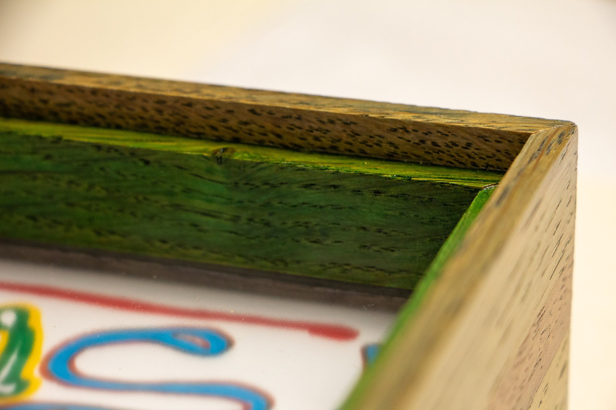 Eclipsing Gravitas 002 frame: angled corner close-up of the frame profile, showing the olive-green toned oak, the vivid green inner construction, clean miter joinery, and the artwork blurred beneath the glazing.