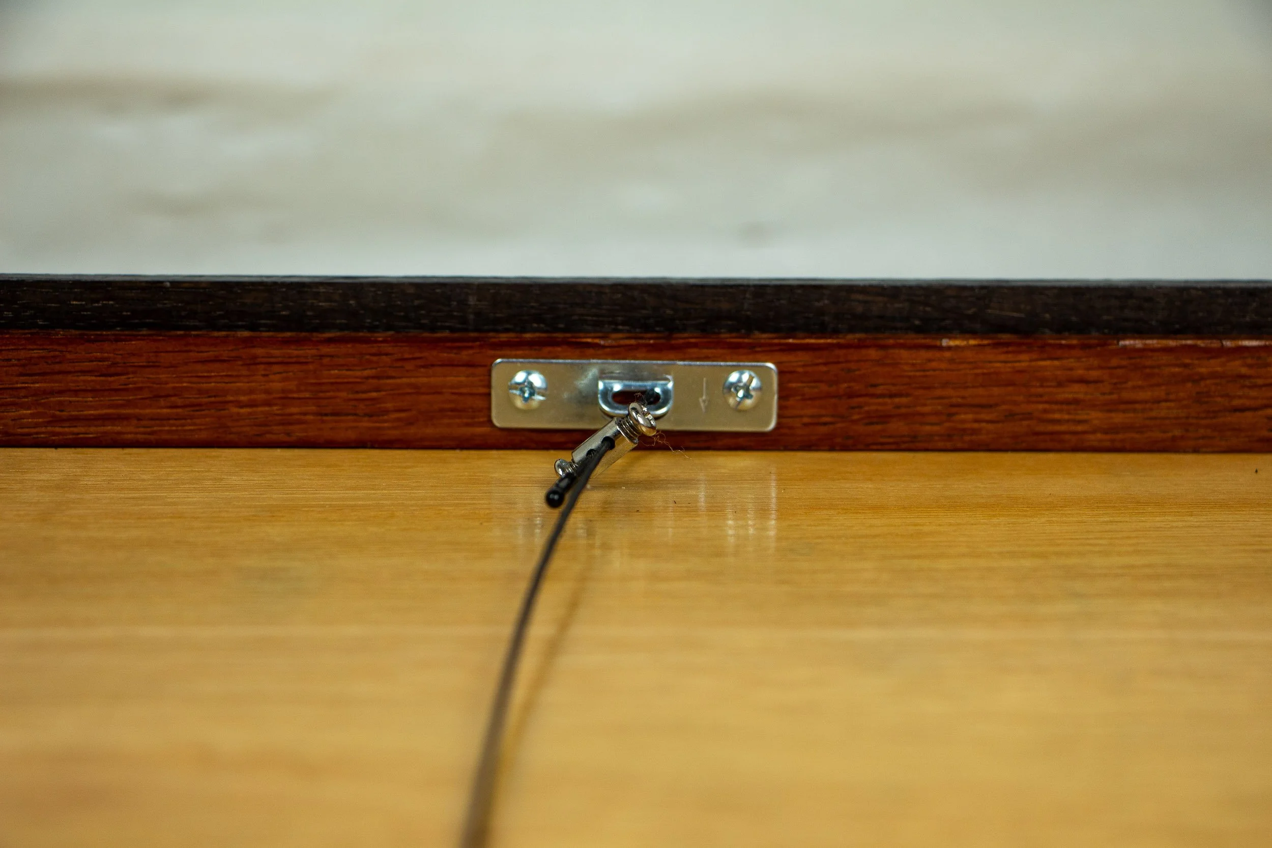 Mêlée of Discovery 005 frame detail: centered close-up of the reverse hanging hardware, showing the metal plate, attached cable, and warm wooden back panel within the frame.