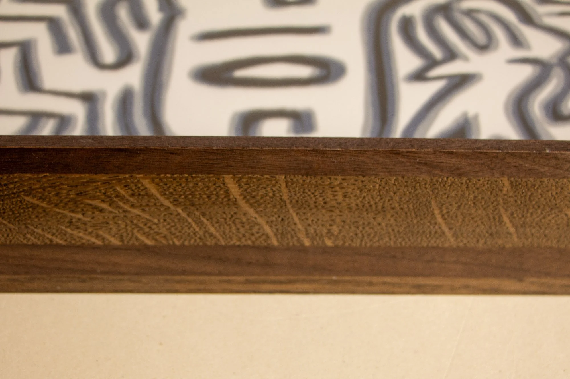 Close-up of a wooden art frame with a decorative wood pattern, with a blurred artwork behind it.