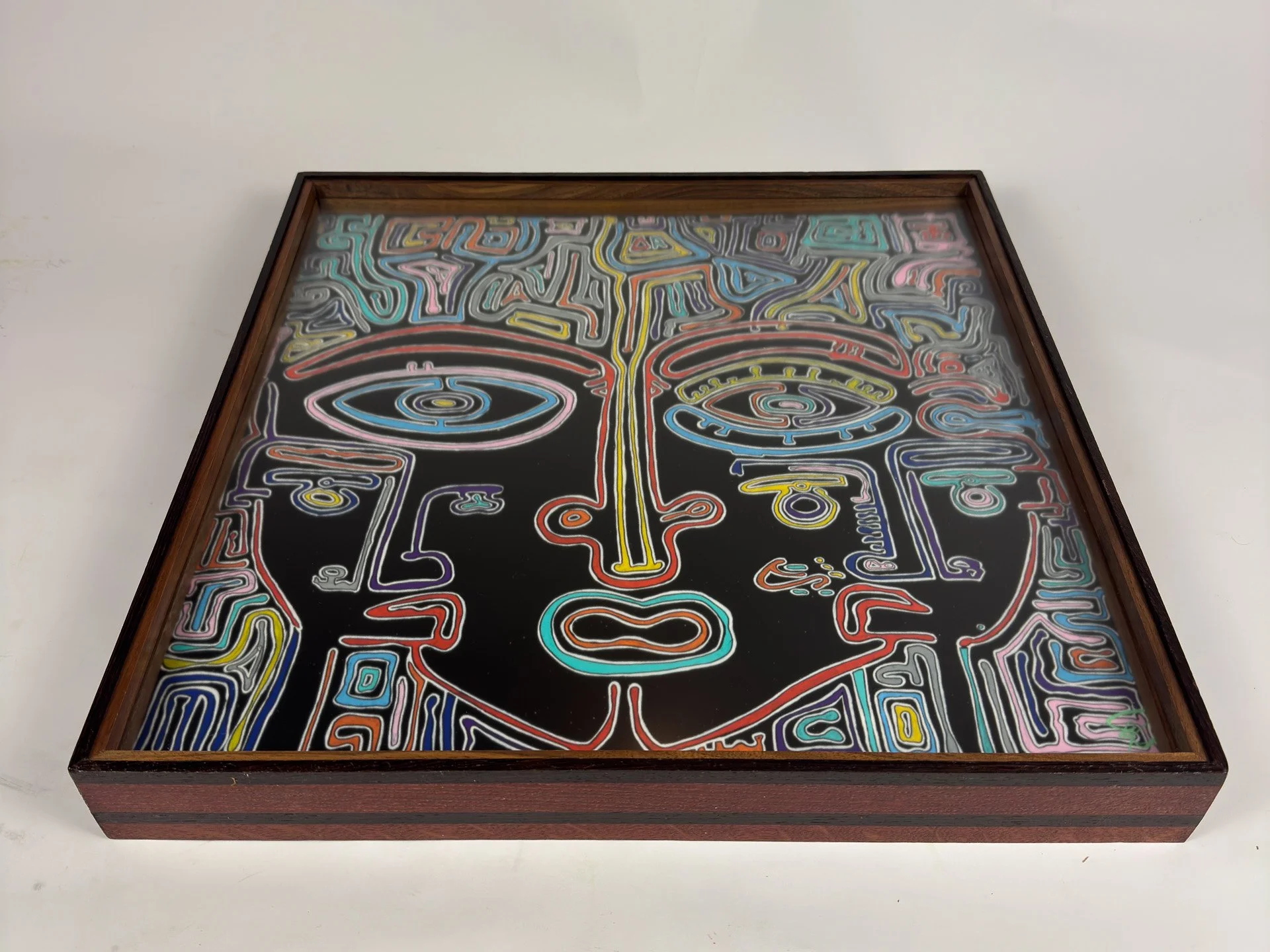 Low-angle view of the artwork set in its custom frame, showing the stylized face behind glazing and the layered construction in dark wengé, warm oak, and walnut, with the frame’s depth giving the piece weight and presence.