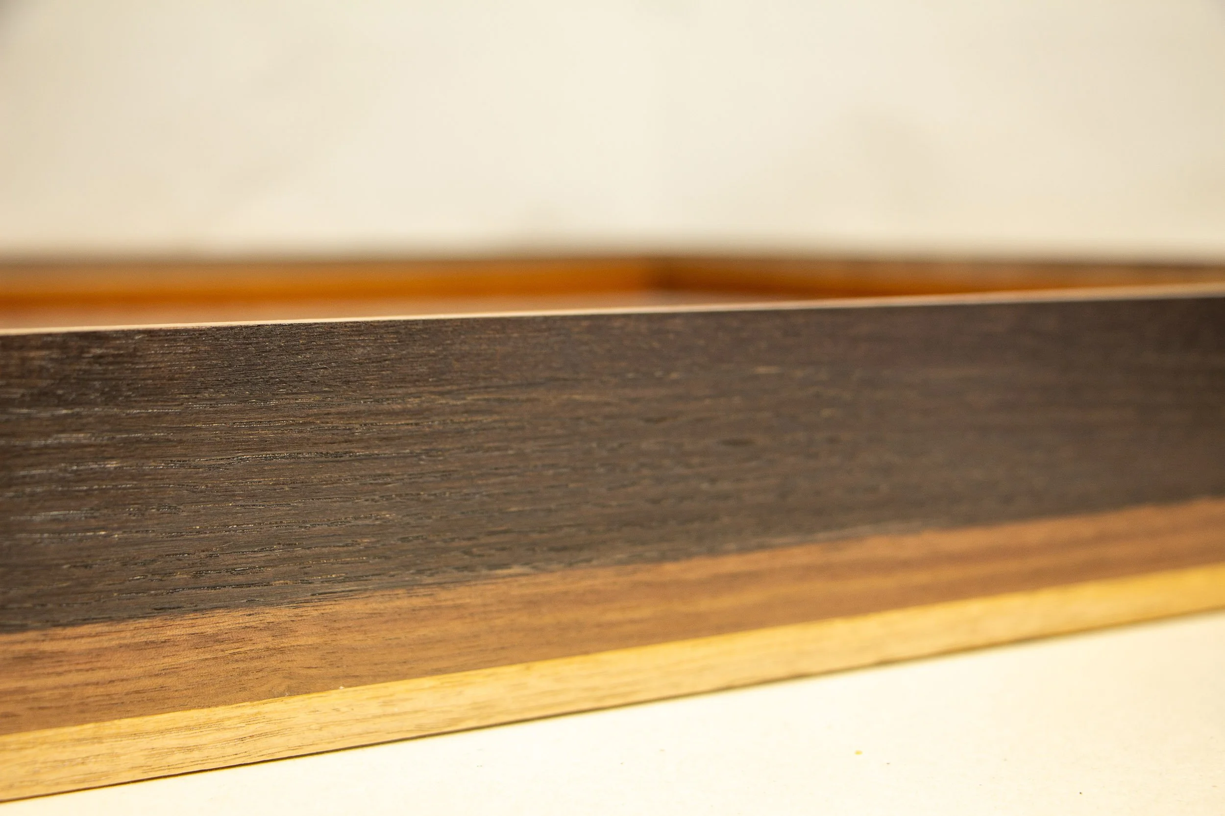 Mêlée of Discovery 004 frame: side profile detail of the exterior, emphasizing the dark upper band and the softer transition through walnut into the light oak base.