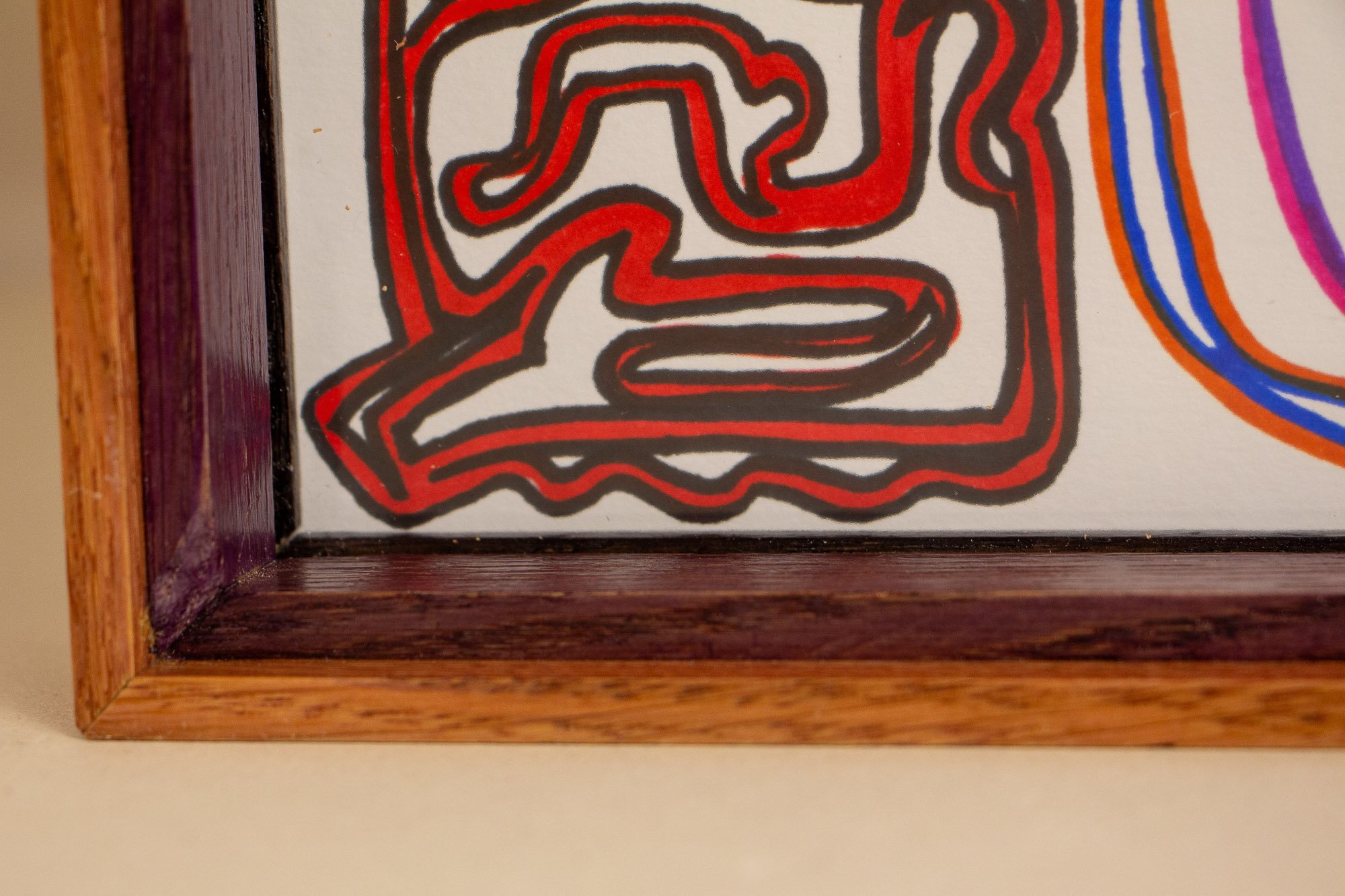 Eclipsing Gravitas 003 frame: lower corner detail from the front, showing the clean miter joinery, the warm pink-orange outer frame, the dark purple inner lip, and a red abstract form meeting the frame edge beneath the glass.