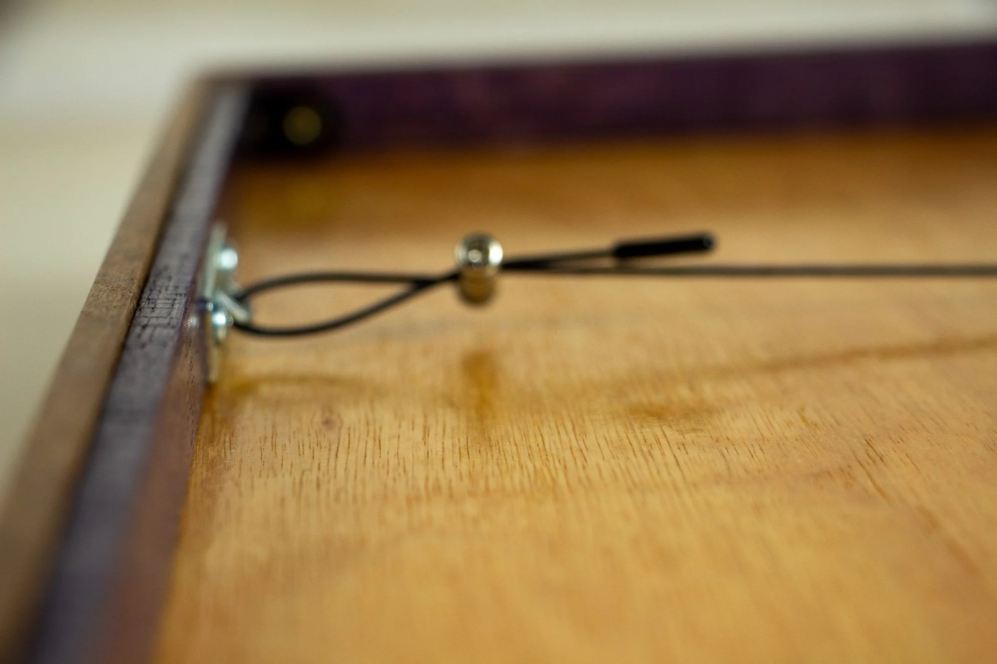 Unseen Signifiers 004 frame detail: reverse close-up of the hanging system from the side, showing the black steel cable, metal fastener, and the warm-toned plywood backing.
