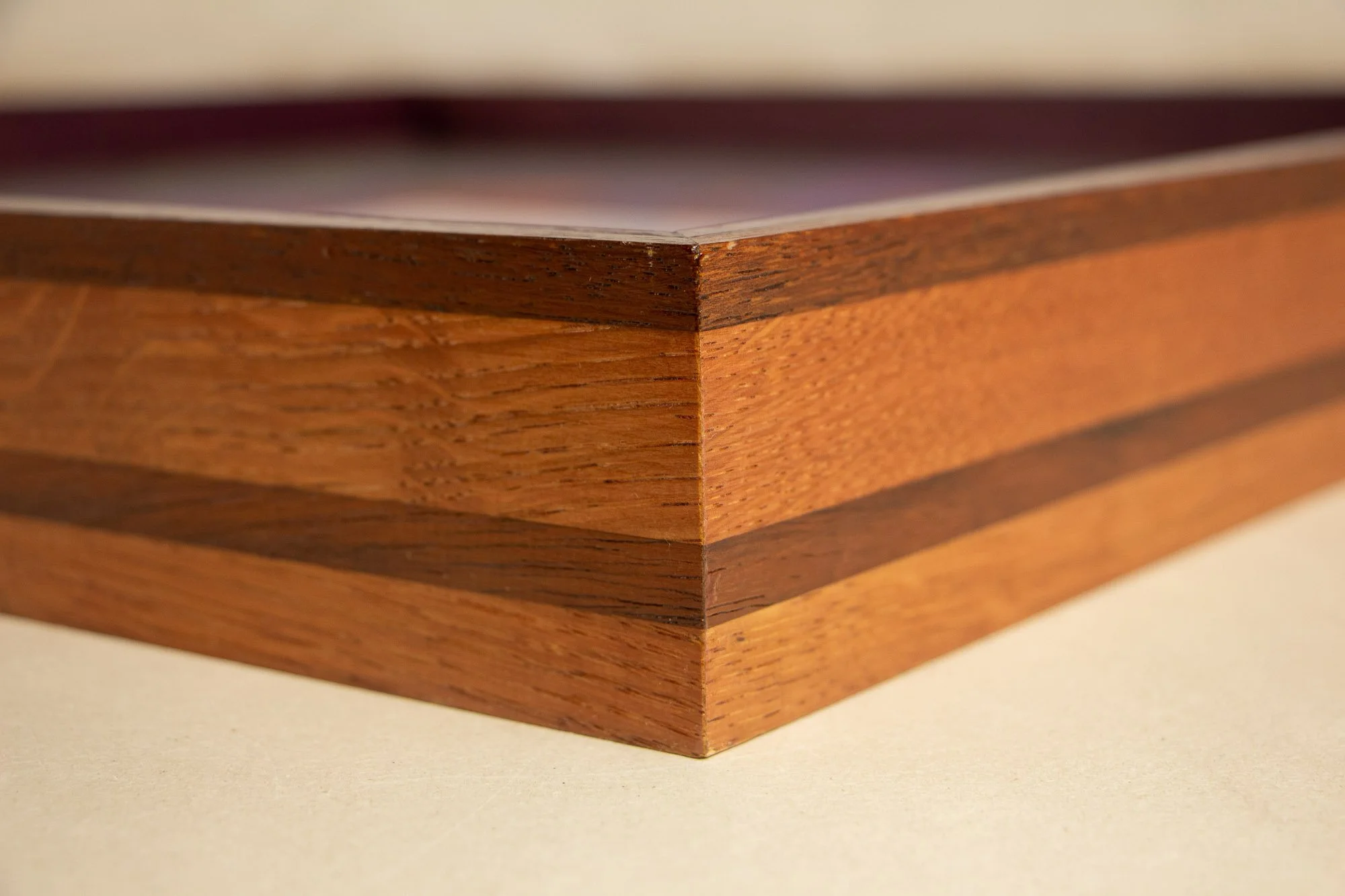 Unseen Signifiers 005 frame detail: angled corner close-up of the layered wooden frame, showing the clear glazing edge, a dark mahogany top band, broad honey-toned oak body, and a darker horizontal inlay running through the profile. mahogany, oak and