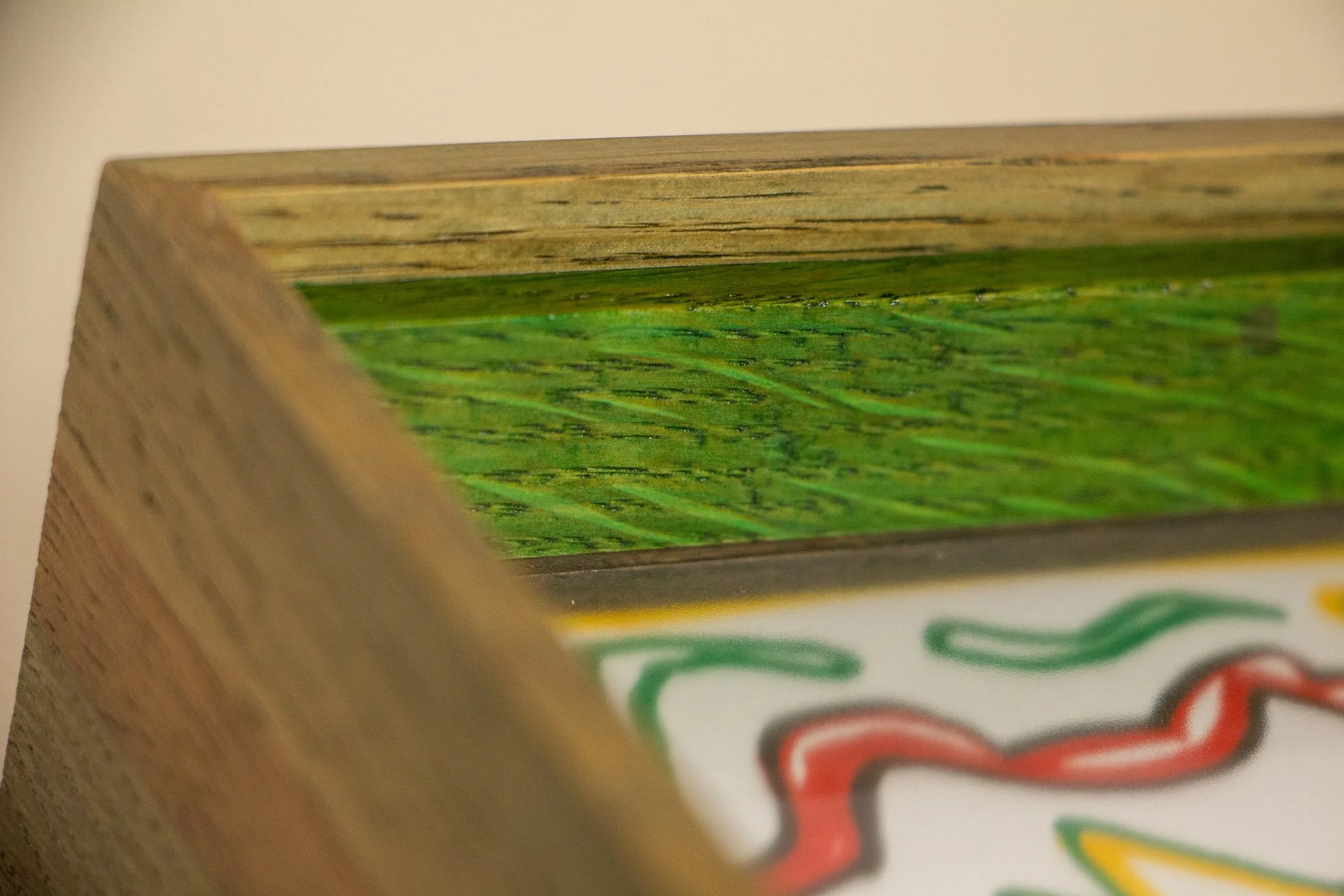 Eclipsing Gravitas 002 frame: macro corner detail showing the layered oak frame with an olive-green toned outer face, a vivid green inner lip, visible natural grain, and the multicolored drawing softly receding beneath glass.