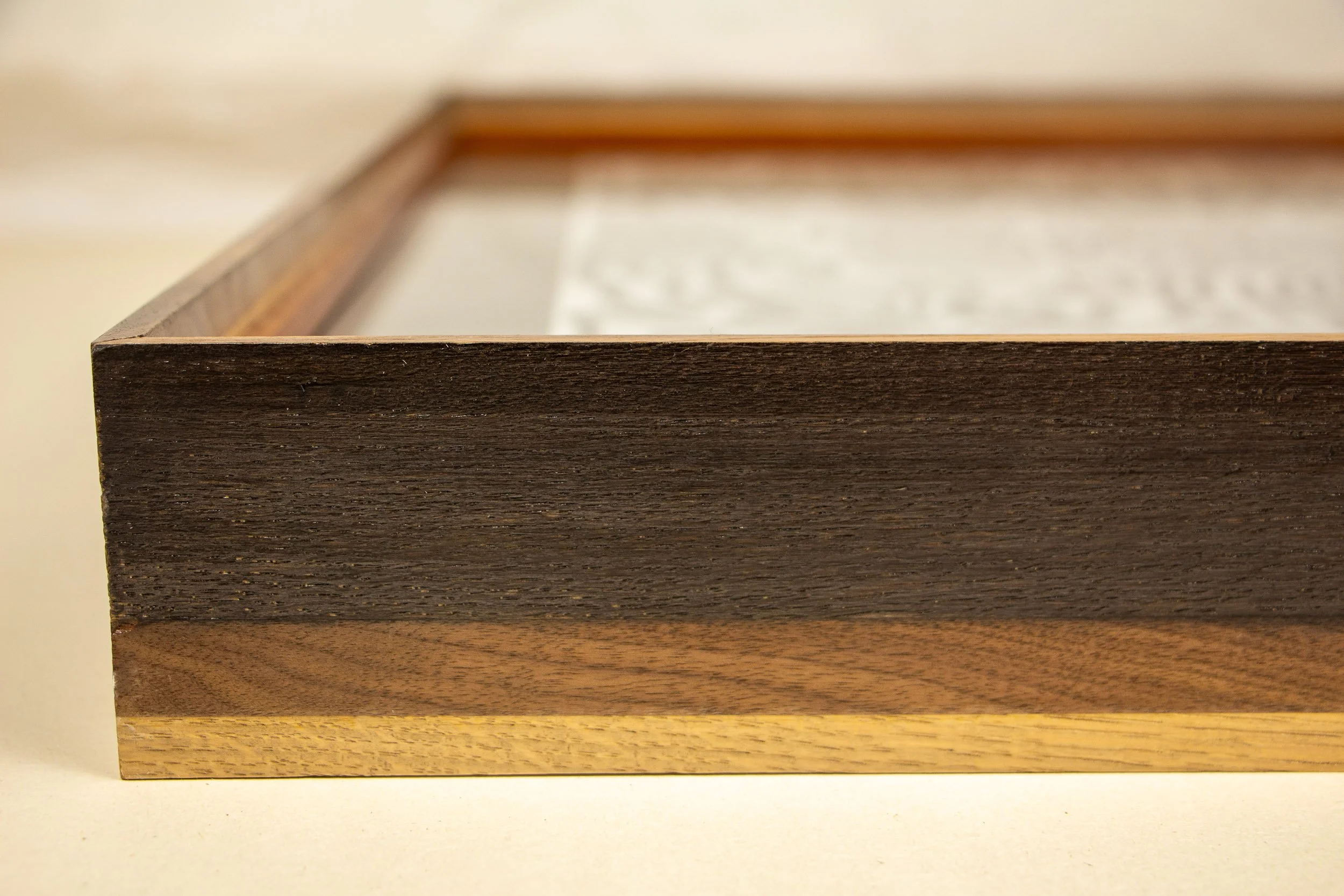 Mêlée of Discovery 002 frame: frontal profile detail of the frame, showing a dark upper band, a warmer walnut middle layer, and a light lower edge arranged in a clear horizontal sequence.