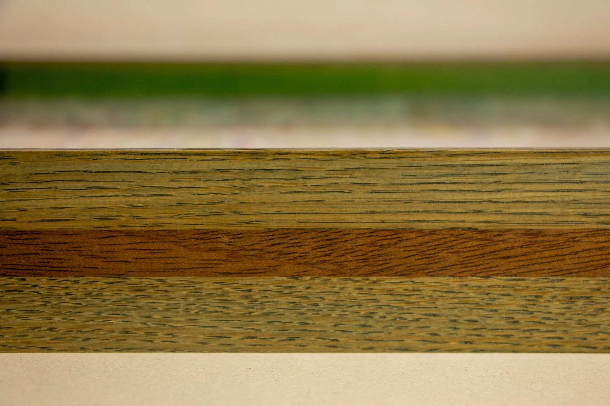 Eclipsing Gravitas 002 frame: frontal macro of the layered frame profile with olive-green toned oak and a warm central mahogany band, showing the low-profile construction and the artwork softly receding in the background.