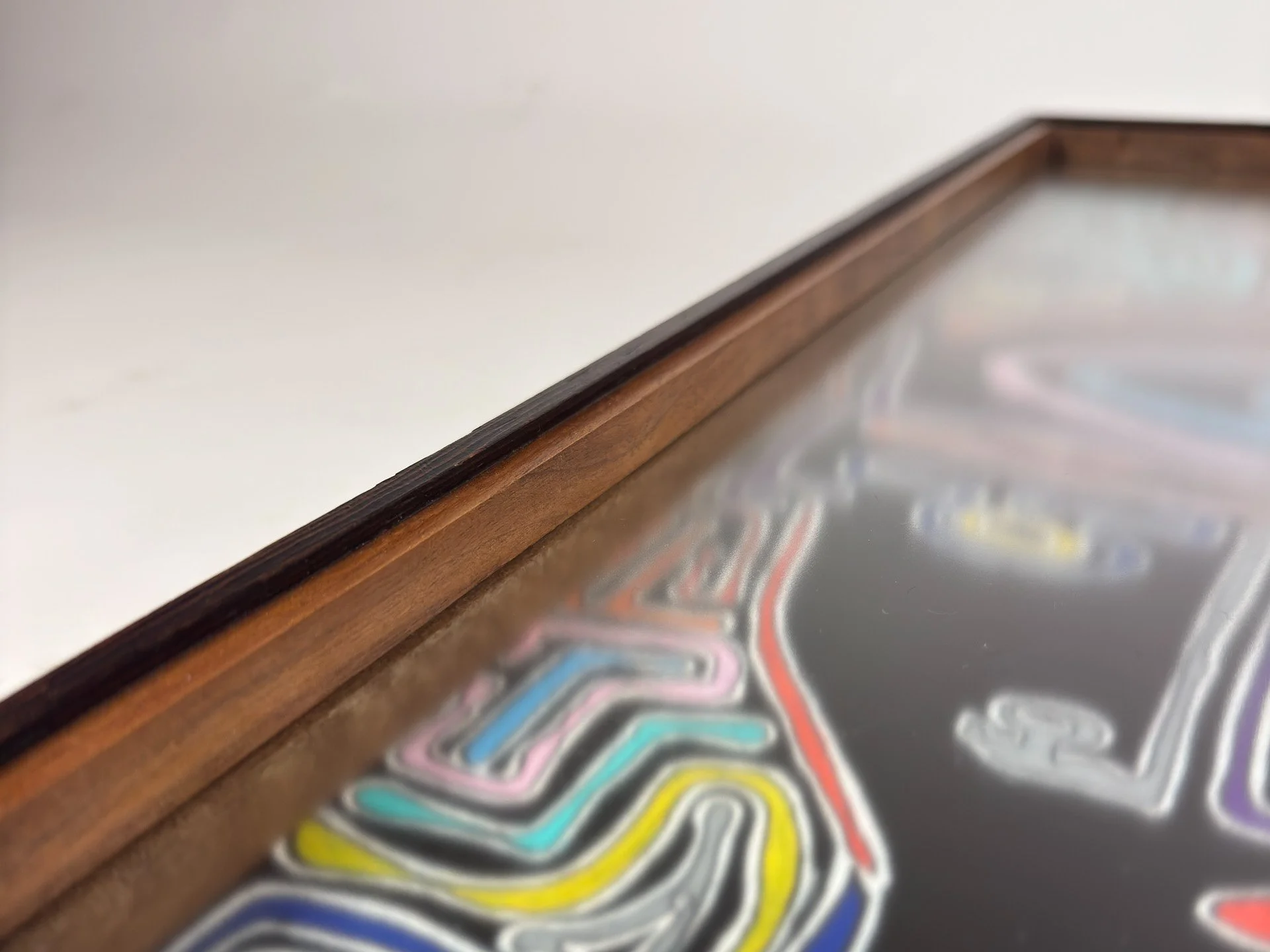 Angled detail along the frame edge, showing the dark outer structure and warm inner walnut line drawing the eye inward, while blurred maze-like lines of the artwork appear beneath the glazing.