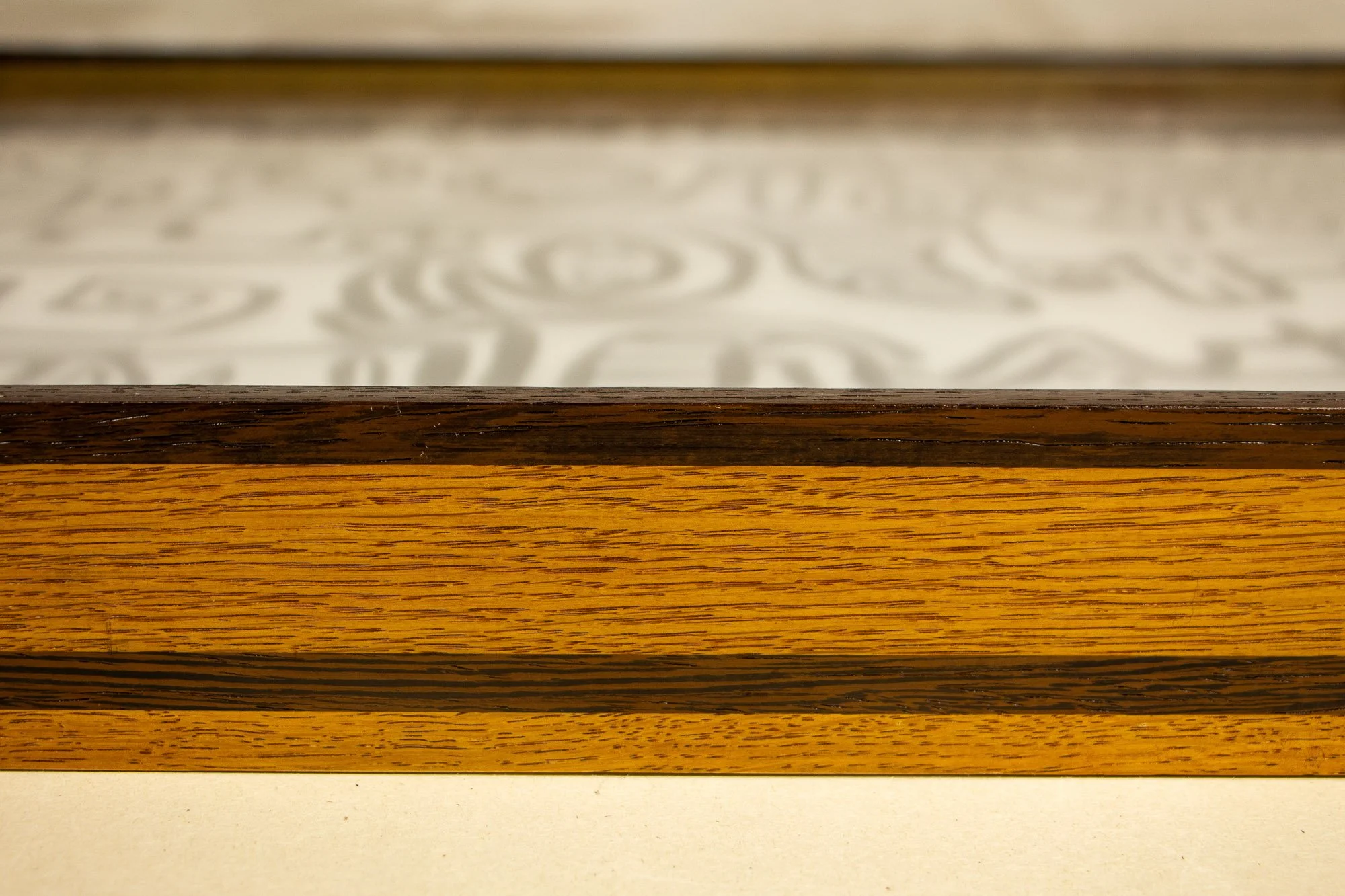 Elusive Cocoon 004 frame: low frontal detail of the frame edge with dark wengé upper rail, broad yellow-toned oak face, and darker central strip beneath the glass-covered drawing.